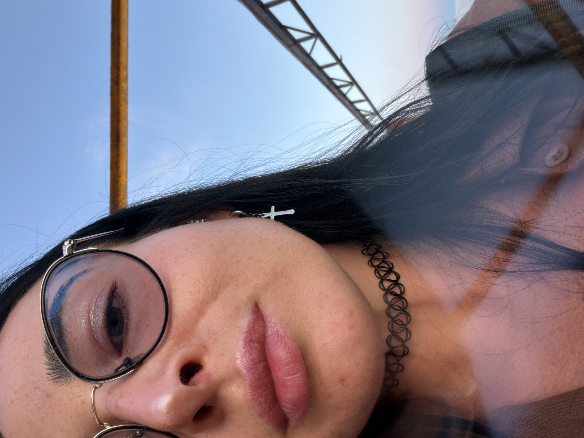 A close-up, chest-up, eye-level, medium outdoor shot shows a young woman with long, dark hair and dark eyebrows. She is wearing round, wire-rimmed glasses with thin gold frames. Her left eye is visible, with a subtle shimmer of eyeshadow. She has full lips with a slight sheen. Around her neck, she wears a black, stretchy tattoo choker. A small silver cross earring hangs from her left earlobe. Her hair is pulled back from her face, but strands of dark hair fall forward. The background consists of a clear blue sky with a thin yellow horizontal bar and a triangular metal structure on the right. A blurred reflection of the photographer is visible on the left side of the image. The lighting suggests it is daytime, possibly late afternoon due to the soft light.