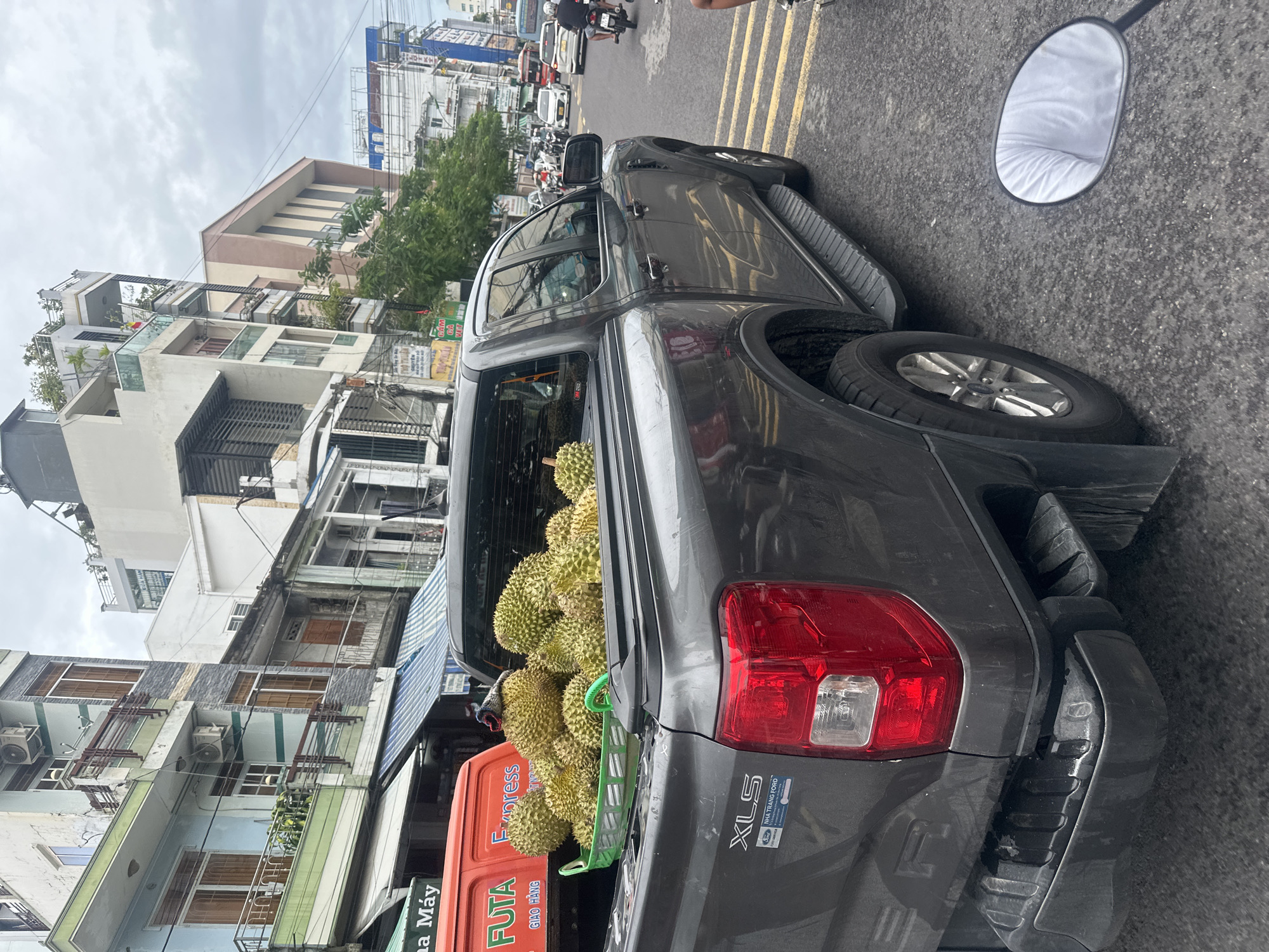 The image depicts a gray pickup truck, parked on a city street next to a building that has "Nhà Máy" on a sign, likely in Nha Trang Ward, Vietnam. The back of the truck is filled with durian fruit. The surrounding area includes other buildings, vehicles (including a scooter), and road markings.