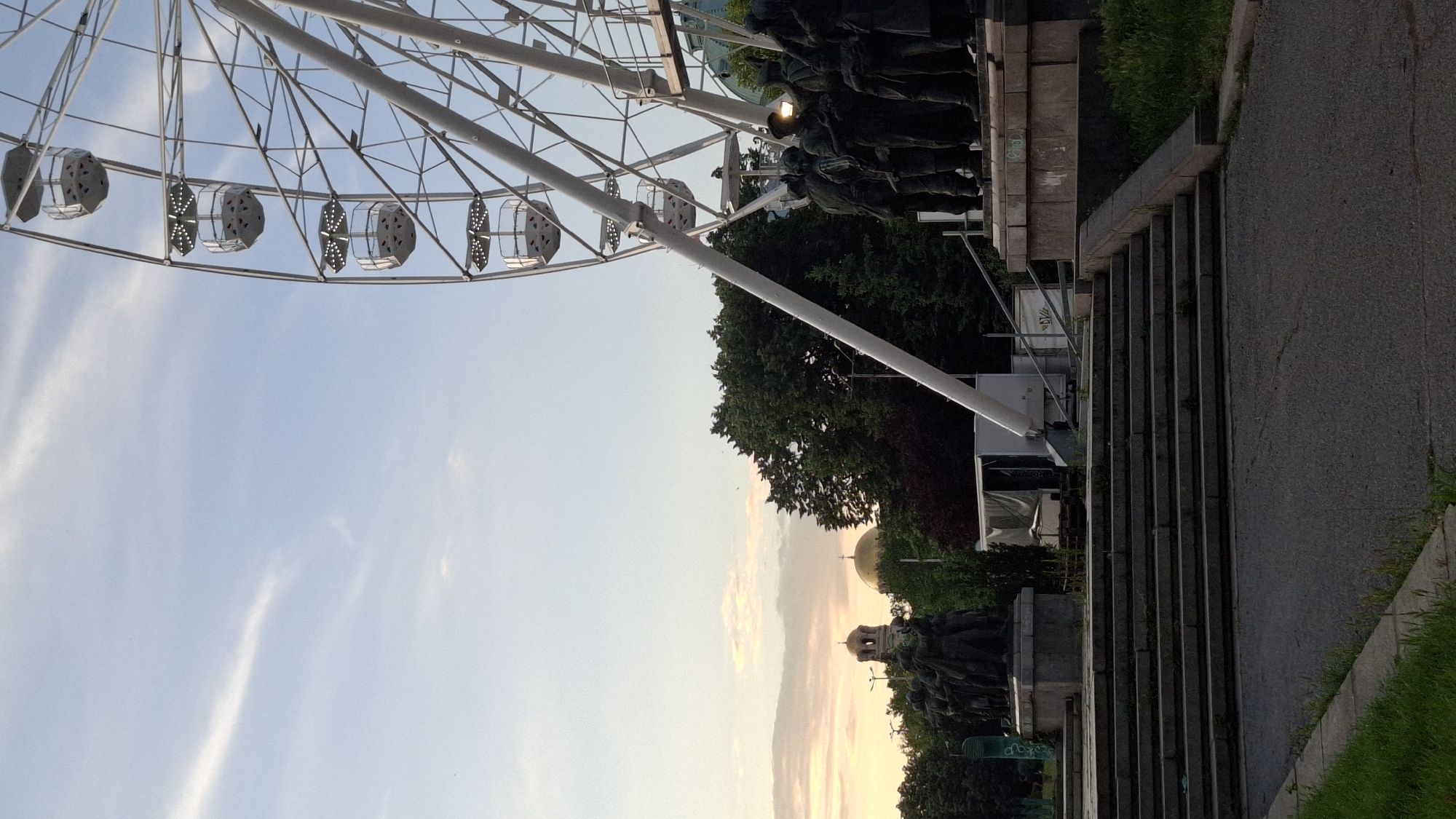 The image captures a scene in Sofia, Bulgaria, featuring the Monument to the Soviet Army in the foreground, with a Ferris wheel and the golden domes of a cathedral in the background.
In the foreground, a wide, gray stone staircase leads up to a plaza. The plaza features a prominent monument, the Monument to the Soviet Army, depicting a group of Soviet soldiers in bronze. The soldiers are depicted standing resolutely, with one soldier appearing to point into the distance. Behind the monument, a white trailer or structure with a sloped roof is visible.
Dominating the right side of the image is a large, white Ferris wheel, its structure and several gondolas clearly visible against the sky. The gondolas are white and have a hexagonal shape.
In the background, lush green trees form a dense canopy. Peeking through the trees are the golden domes of a cathedral, likely the Saint Alexander Nevsky Cathedral, a prominent landmark in Sofia. The sky above is a soft blue with scattered wispy clouds, and the lighting suggests it is either early morning or late afternoon, with a warm glow on the horizon. The overall impression is a blend of historical monuments, urban recreation, and architectural beauty within the city of Sofia.