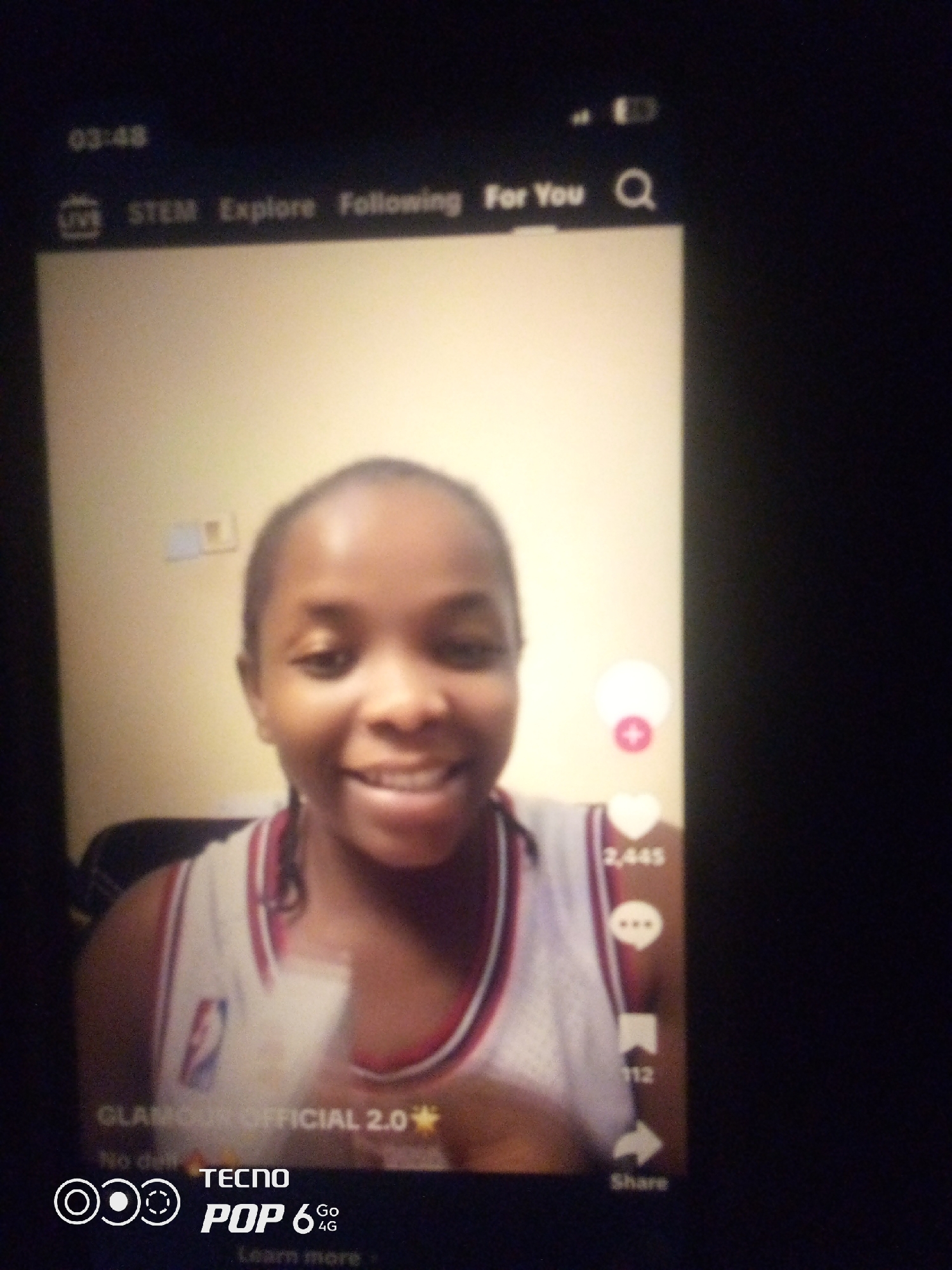 The image displays a smartphone screen showing a short-form video. A young Black female with braided hair is smiling at the camera, wearing a white and red-trimmed tank top with an NBA logo. The video interface shows icons for "LIVE," "STEM," "Explore," "Following," and "For You." A search icon is visible. On the right side of the screen, there are icons for likes, comments, and shares, with "2,445" indicating the number of likes. The bottom of the screen displays "GLAMOUR OFFICIAL 2.0," "TECNO POP 6 Go 4G," and "Learn more." The background appears to be an indoor setting with a light-colored wall. The image quality is low, with a blurred appearance. There is no specific information within the image to indicate a location in Howayi, Nigeria.