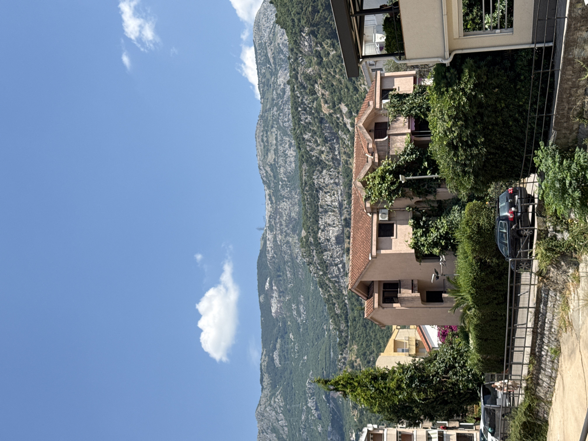 A landscape scene in Budva, Montenegro, captures a clear blue sky with sparse white clouds above a verdant mountain range. In the foreground, a building with a terracotta roof is visible, covered in ivy. Adjacent to it, a modern building with balconies stands. A walkway with a metal railing borders a lower area, with a few cars parked along the road. A woman is walking on the sidewalk, and lush green bushes and trees are abundant throughout the scene, indicating a sunny day with warm weather.