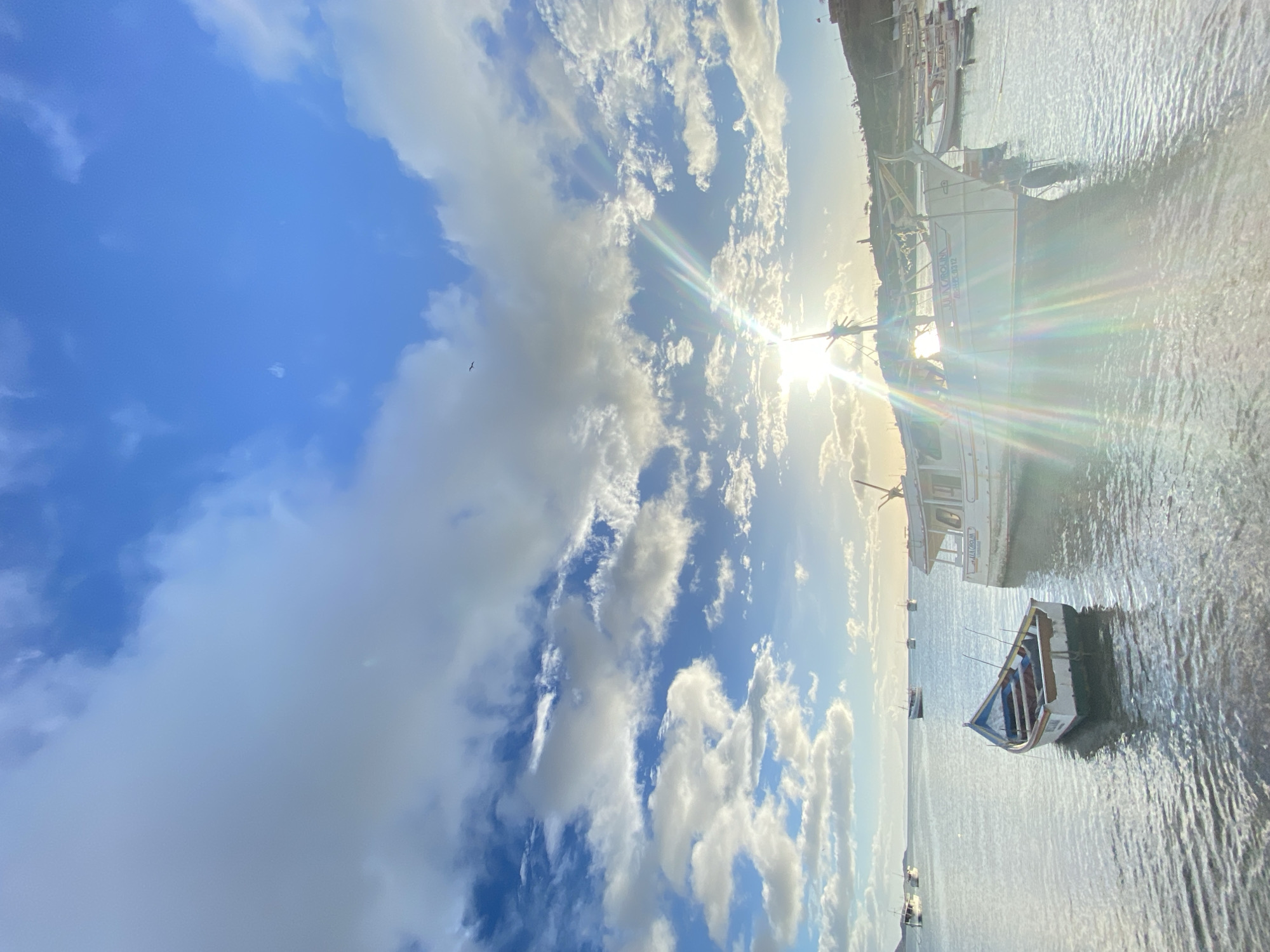 The image depicts a daytime scene in Juan Griego, Venezuela. The sky is blue with scattered white clouds, with the sun shining brightly through them, creating a starburst effect. The foreground features a calm body of water, possibly a harbor or bay, reflecting the sunlight. Several boats are present, including a small rowboat in the lower right and larger vessels docked nearby. The boats are of different sizes and designs. The overall environment suggests a coastal setting.