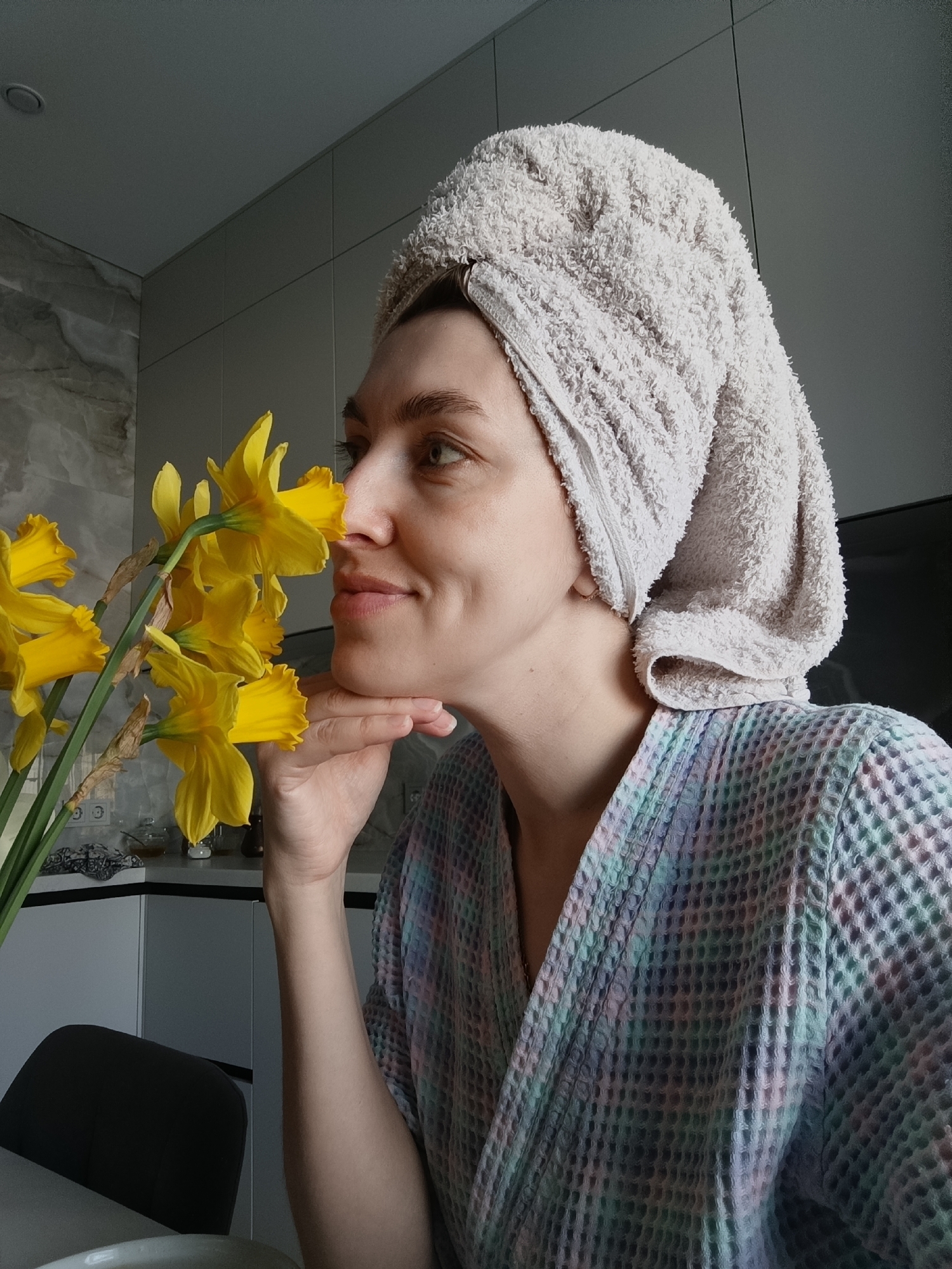 A woman with a towel wrapped around her head is smelling a bunch of yellow daffodils. She is wearing a waffle weave bathrobe. She is holding her chin with her left hand. The background shows a kitchen with light gray cabinets and marble backsplash. A black chair is visible in the foreground. Based on available information the city may be related to the images's origin.