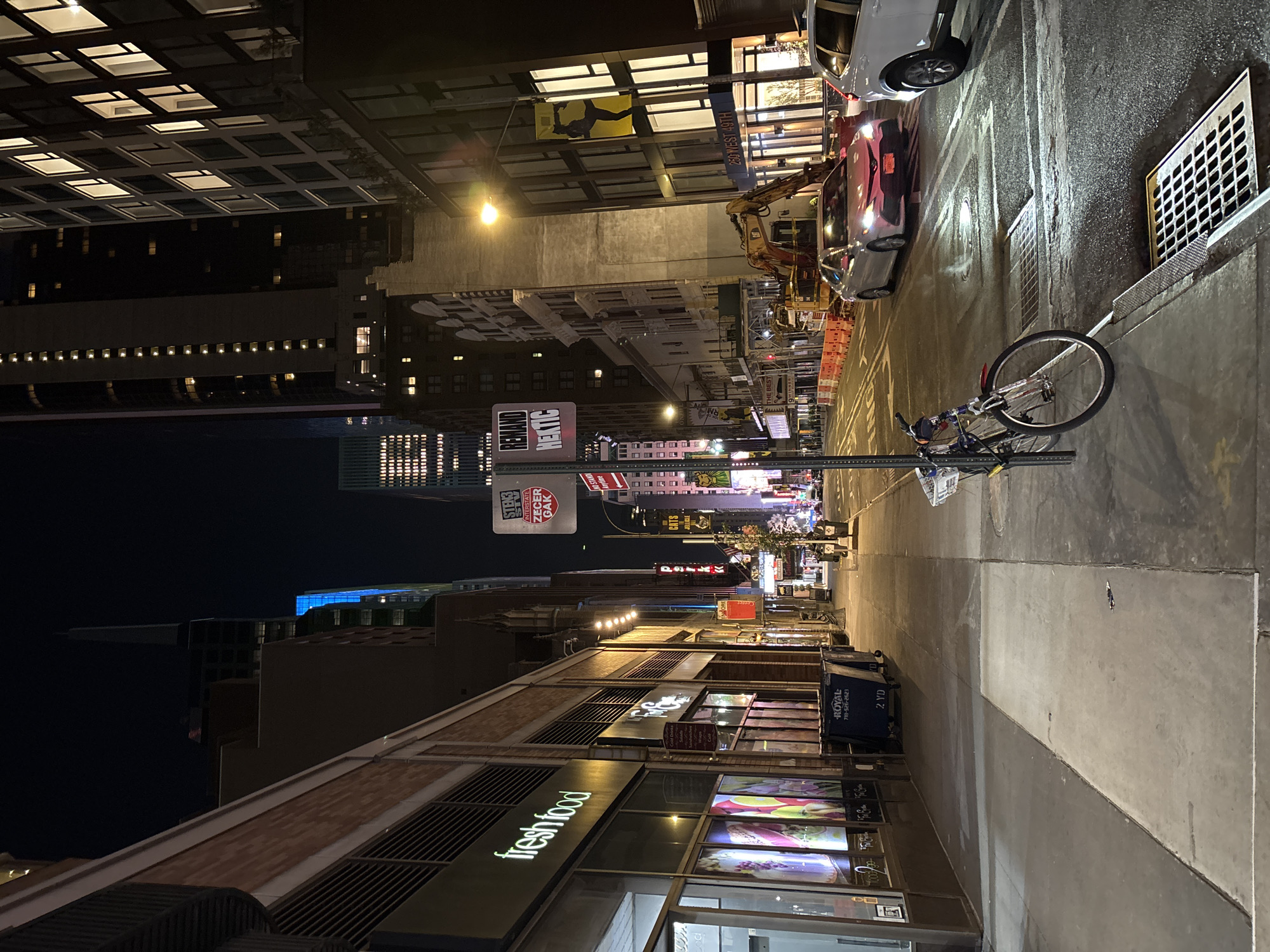 This is a nighttime photograph depicting a vibrant urban street scene, likely in New York City, given the "250 WEST 49TH" sign. The street and sidewalk are illuminated by numerous artificial lights, casting shadows and reflections.
In the foreground, a "freshfood" store occupies the ground floor of a building on the left, with brightly lit display windows showcasing various food items. A blue "ROYAL" trash or recycling bin, marked with "718-526-2623" and "2 YD", stands on the sidewalk. The pavement shows signs of wear and repair.
Moving into the midground, a bicycle is parked against a metal pole on the sidewalk. Further down the street, an excavator or construction vehicle is visible next to a parked red car, which itself is behind a white SUV, suggesting construction or roadwork. Traffic cones are also present. The sidewalk features a large metal grate. Numerous buildings line both sides of the street, with many windows lit up, some with distinctive patterns.
In the background, the street recedes into a perspective of brightly lit signs and building facades, typical of a busy commercial area. The night sky is dark, contrasting with the illuminated cityscape.
Visible text includes:
- "freshfood"
- "ROYAL" with phone number "718-526-2623" and "2 YD"
- "Allstate" (partially visible logo) and "ZEGER GAK"
- "NO STAND Anytime" (street sign)
- "REMAND HEKTIC" (sign on a pole)
- "park" (small sign)
- "250 WEST 49TH" (building address)
- "CAT" (on the construction vehicle)
- Distant signs like "THE BEST," "CATS," and "JUICE" are also faintly discernible.
The scene captures the dynamic, bustling atmosphere of a city at night, with a mix of commerce, transportation, and residential/office buildings. The clear weather allows for sharp visibility of the street's details and distant lights.