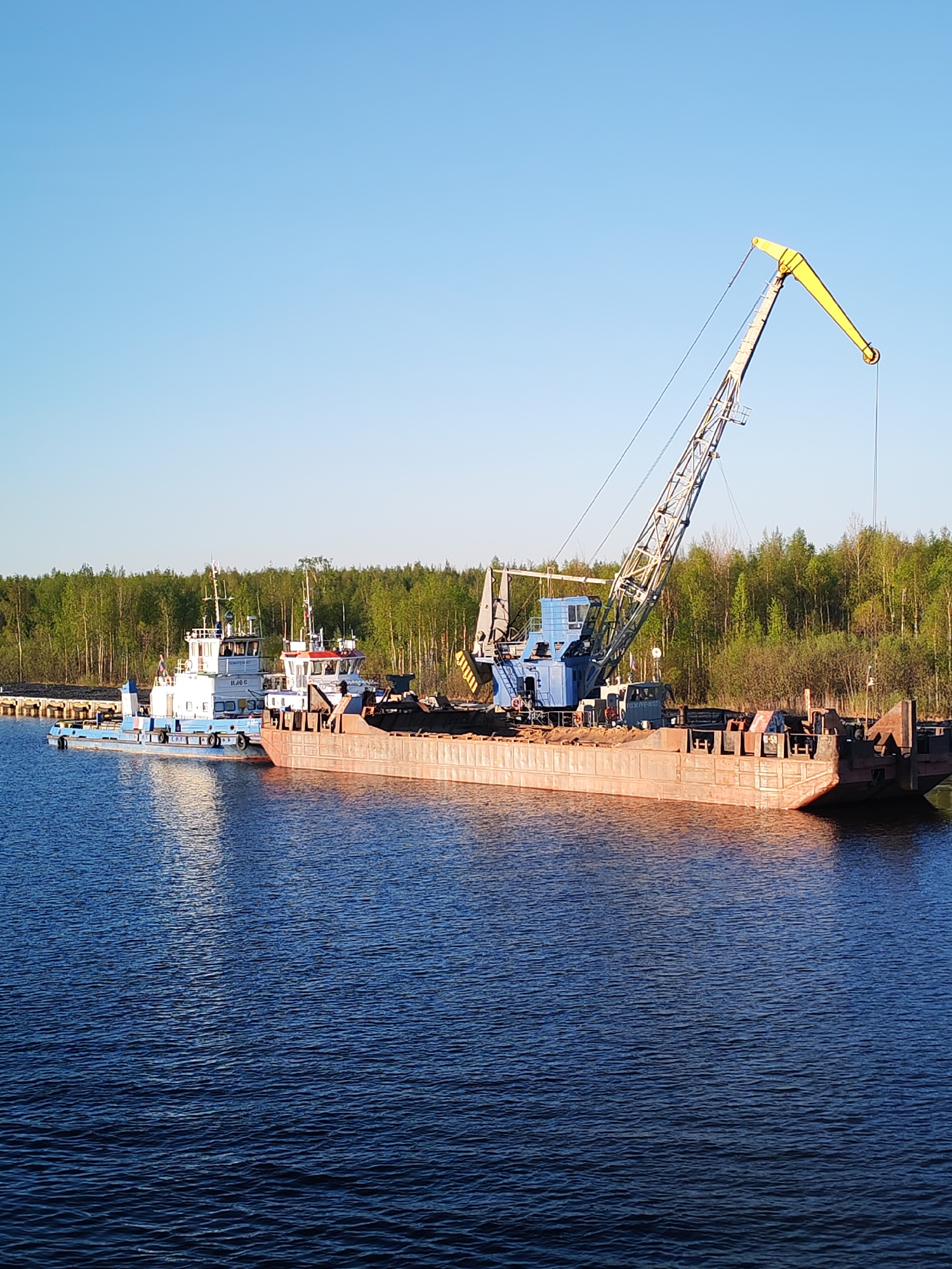 The image depicts a tugboat connected to a barge equipped with a crane in the city of Ozernoe Ust'e, Russia. The crane is positioned on the barge with a yellow tip. The tugboat is painted white and blue, while the barge appears to be brown or rust-colored. The background consists of a forested area, and the scene is set on a body of water under a clear blue sky. The water surface has subtle ripples. The overall scene suggests a waterway with industrial or cargo handling activity.