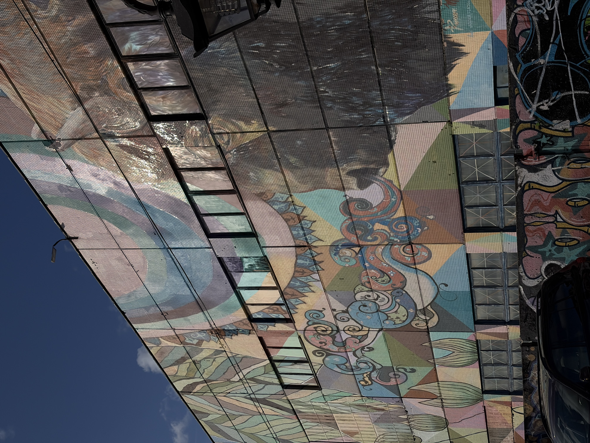 This is a photograph of a large building in Minsk, Belarus, featuring a vibrant and colorful mural on its facade. The mural appears to be made of mosaic tiles and depicts abstract shapes, swirling patterns, and stylized natural elements like leaves and possibly floral motifs. The upper portion of the mural also seems to incorporate a large, somewhat abstract portrait or face rendered in muted tones of brown, beige, and gray, with a dark, feathery texture. Several rectangular windows are visible, breaking up the mural. A street lamp is mounted on the top edge of the building, and a black car is parked in front of the building at the bottom of the frame. Below the mosaic mural, a section of the building is covered in graffiti, adding another layer of urban art to the scene. The sky above is a clear, deep blue with a few wispy clouds. The overall impression is one of urban decay juxtaposed with bold, artistic expression.