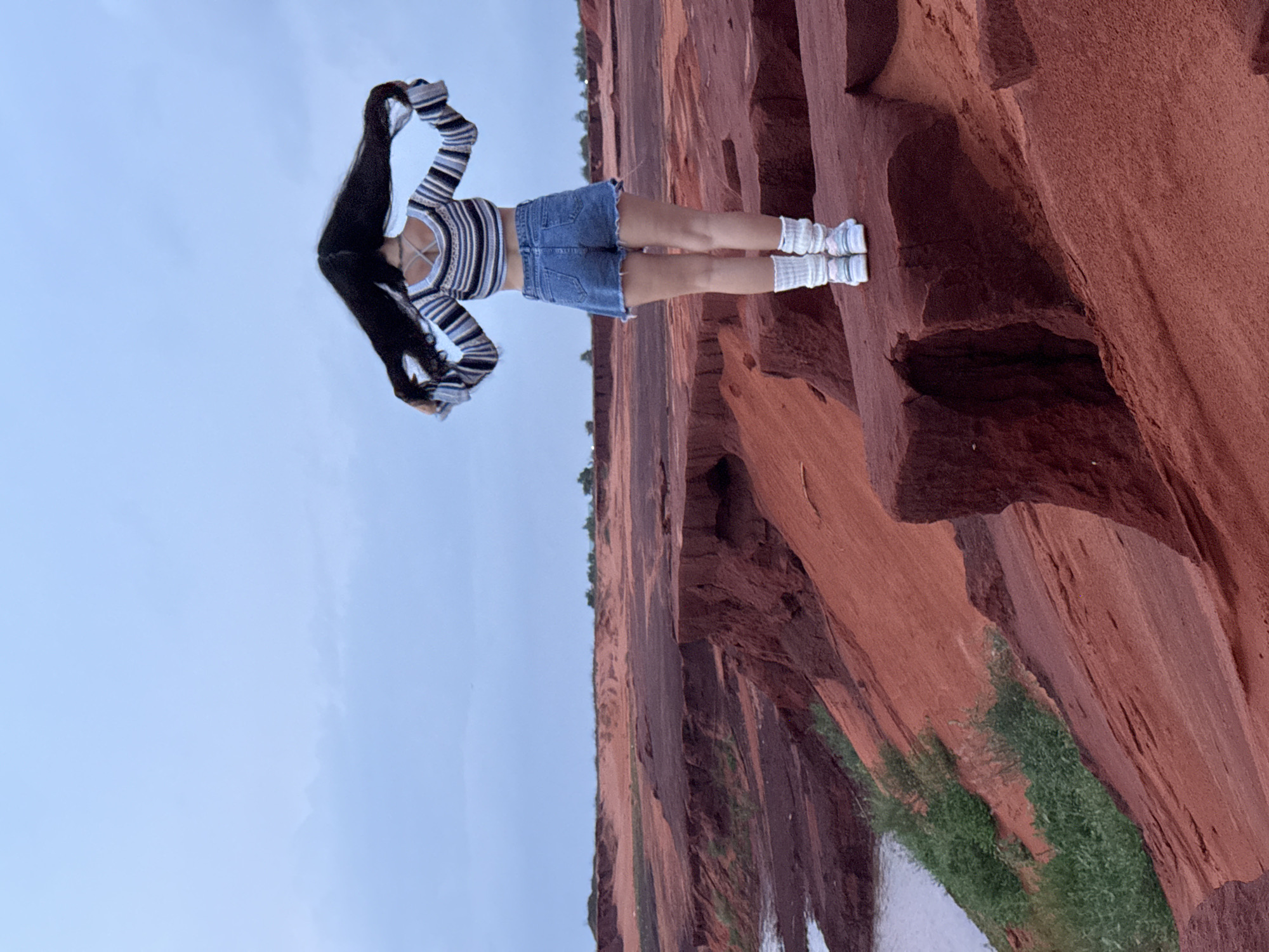 A person stands in a unique natural landscape dominated by towering reddish-brown rock formations. The individual, with long, dark hair flowing upwards, is dressed in a horizontally striped long-sleeve crop top, a denim mini skirt, white scrunchy socks, and light-colored shoes. Their arms are raised above their head, contributing to a dynamic pose.
The background features steep, rugged cliffs or canyon walls with varied textures and shades of red and brown. Sparse green vegetation grows at the base of these formations and along the water's edge. A narrow body of water, likely a stream or river, winds through the canyon floor, reflecting the clear, light blue sky above. The scene appears to be set during daytime, with soft lighting that suggests either morning or late afternoon. The distinctive red rock and water features are characteristic of locations such as the Fairy Stream (Suoi Tien) or similar geological sites found in Mui Ne, Vietnam. No text is visible in the image.