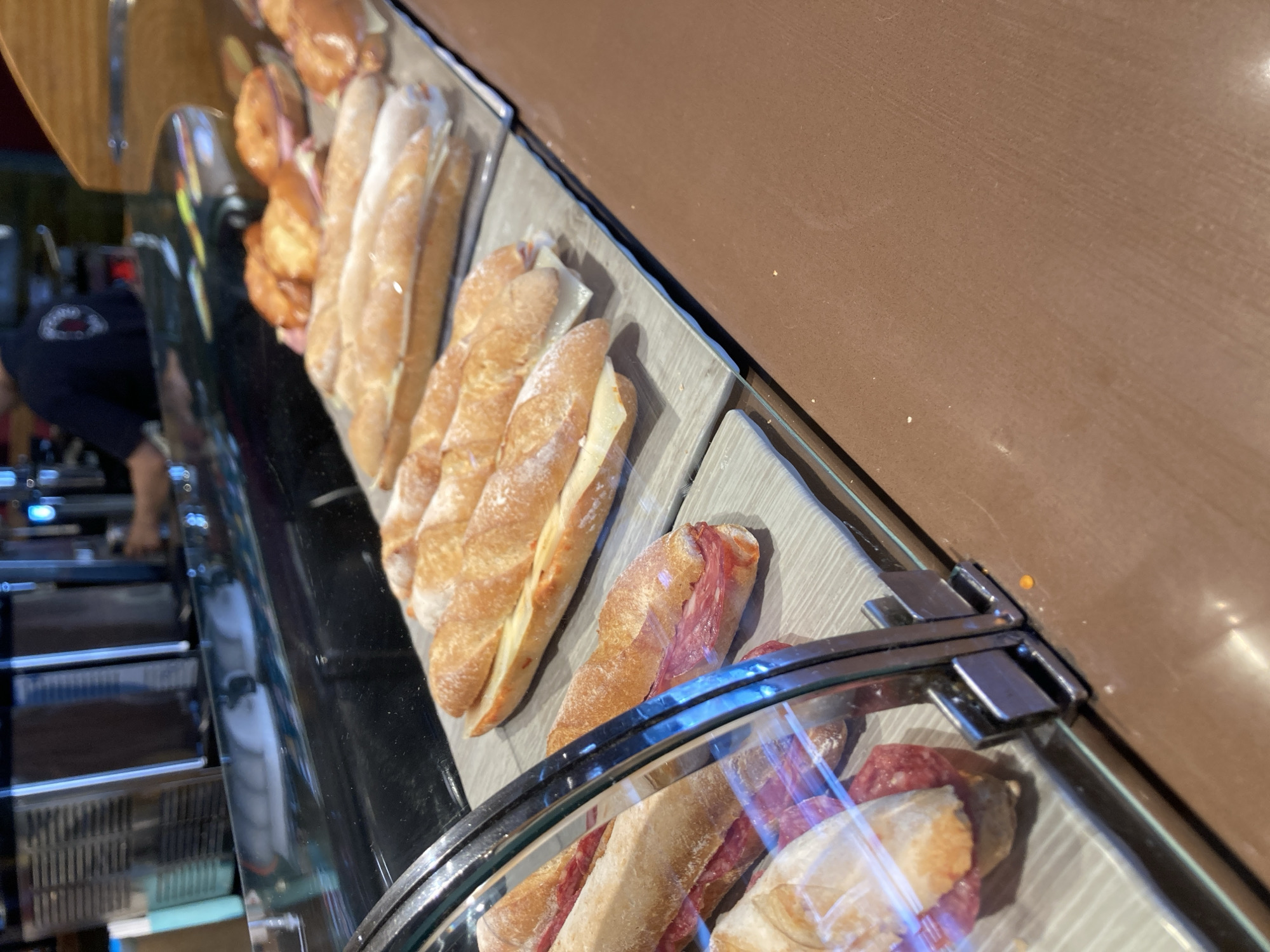 The image shows a display of sandwiches at what appears to be a bakery or cafe. The sandwiches are made with baguettes and filled with cured meats, likely salami, and some appear to have cheese as well. They are arranged on gray display shelves behind a curved glass counter. In the background, a person wearing a dark shirt with a logo is visible, possibly an employee. The scene is indoors, with a warm brown countertop or wall in the background. The lighting suggests it could be daytime. The overall impression is a well-stocked food service establishment. The location is identified as Barcelona, Spain, but there are no specific visual cues to confirm this from the image itself.