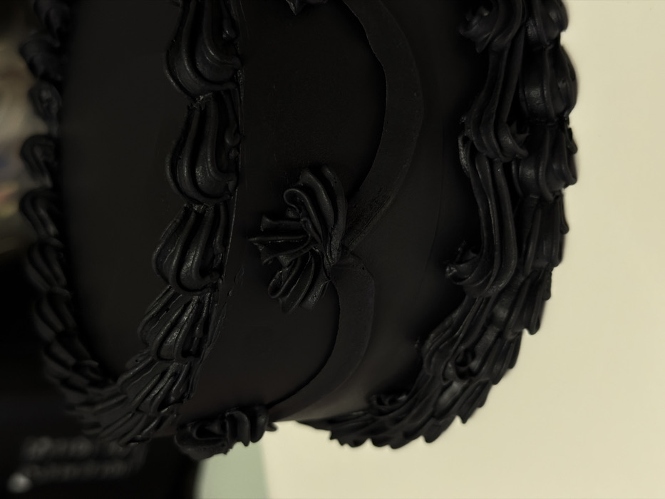 A single-tier, round cake, uniformly covered in smooth, matte black frosting, occupies the majority of the frame, positioned on a light-colored surface, likely a table or cake stand. The cake is elaborately decorated with piped black frosting. A continuous border of stylized scrolls or swags adorns the top edge. The side of the cake features a horizontal band of black frosting, shaped into a draped or looped design, accented by several rosette-like formations. The base of the cake is encircled by a wide, wavy border of textured black frosting. The background is dark and out of focus, providing no discernible details about the broader environment. No individuals, actions, or interactions are visible. The image contains no visual cues, such as landmarks, signage, or distinct architectural styles, to indicate its location in Tel-Aviv, Israel.