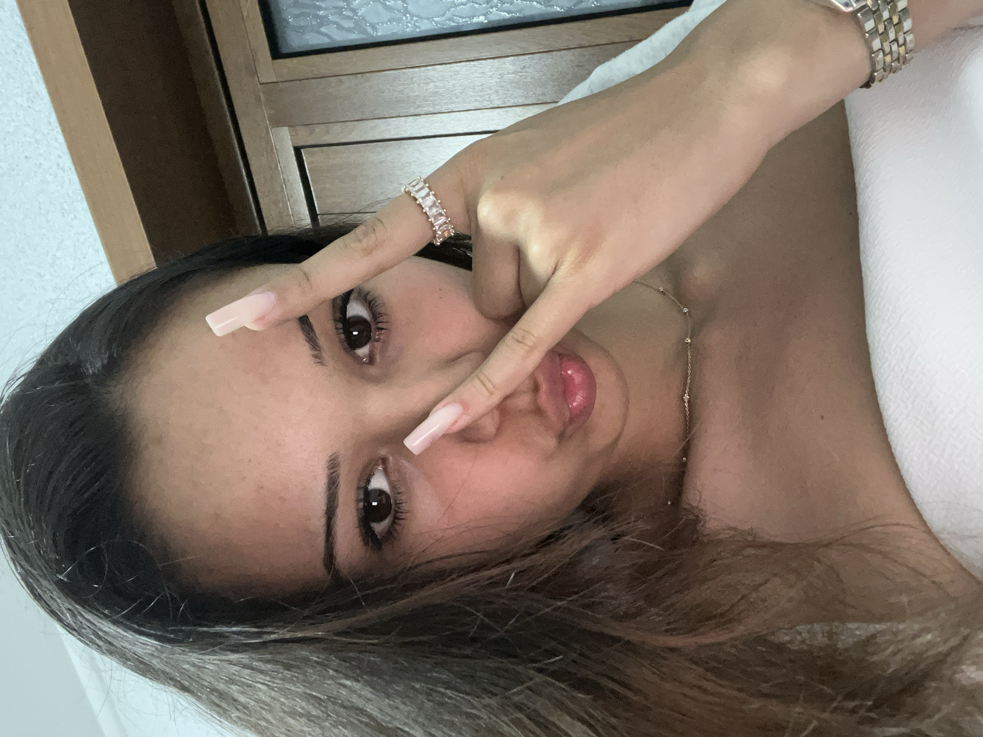 A close-up, high-angle portrait features a young woman lying on her back, taking a selfie. Her head is positioned on the left side of the frame, tilted to the left, giving the impression that she is looking up at the camera. Her dark hair, with lighter brown highlights, flows around her head. She has dark, well-defined eyebrows and long eyelashes. Her left eye is in full view, gazing directly at the camera, while her right eye is partially obscured by her fingers.
She forms a peace sign with her left hand, covering part of her face. Her fingernails are long and manicured with a light pink or nude polish. On her left ring finger, she wears a ring adorned with several rectangular, clear stones. Her lips are slightly parted, revealing a shiny, pinkish hue.
A golden chain necklace with small, spaced-out beads is visible on her neck. On her right wrist, a watch with a silver and gold band is apparent. The background is indistinct, but hints of a wooden surface and a white, possibly textured, fabric can be seen. The overall lighting is soft, suggesting an indoor setting. The woman appears relaxed, with a soft, natural expression.