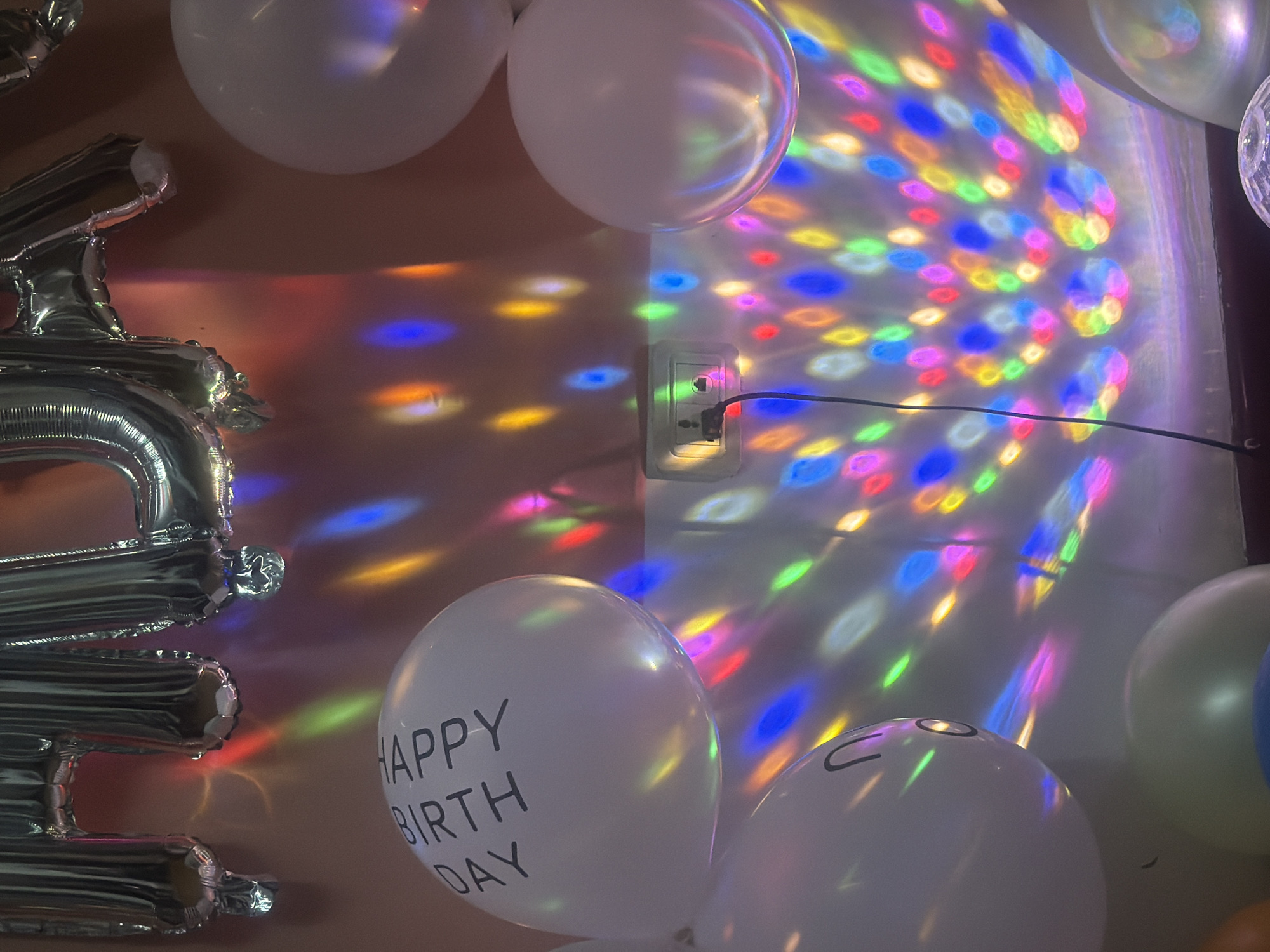 A close-up indoor shot shows a wall partly covered with a pattern of colored lights, next to a standard North American-style electrical outlet with a black cord plugged in. Several white balloons are visible, one with "HAPPY BIRTHDAY" printed on it in black lettering. A silver "2" balloon is also visible on the left. The location is presumed to be Zhongtun, China.