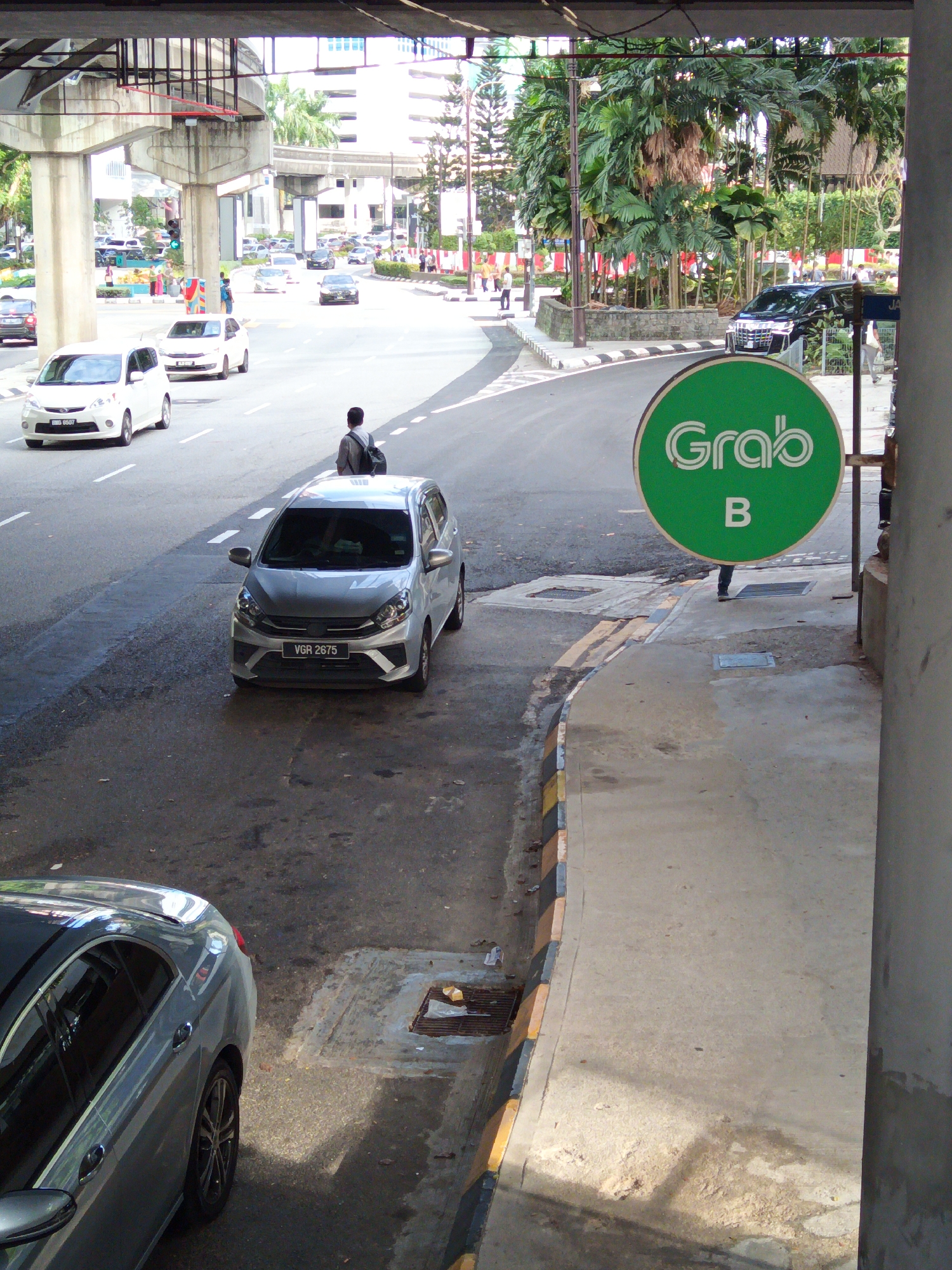 The image captures a street scene in Kuala Lumpur, Malaysia, likely in an urban area with elevated infrastructure. In the foreground, a silver car with the license plate "VGR 2675" is parked on the right side of the road, facing away from the viewer. A person with a backpack is standing behind the car, looking to the right. To the right of the road, a bright green circular sign with "Grab" logo and the letter "B" is mounted on a pole. The background shows a road with several other vehicles, including white cars and a black SUV. Elevated concrete structures, possibly part of a highway or monorail system, are visible overhead. Lush green palm trees and other vegetation line the side of the road, suggesting a tropical environment. The lighting indicates it is daytime, with sunlight casting shadows on the pavement.