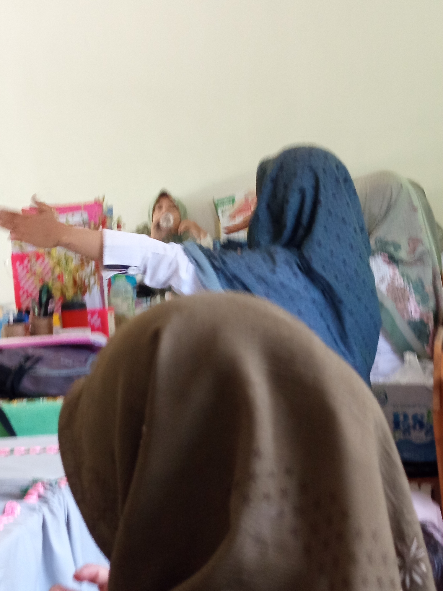 The image depicts an indoor scene with three individuals, two of whom are partially obscured. In the foreground, a person wearing a brown headscarf is facing away from the camera, with their back and shoulder visible. To the left and slightly behind, a person in a white shirt and hijab is reaching out with their left arm, with their hand extended forward. This individual appears to be a woman. Further in the background, a third person, possibly a child, is visible from the shoulders up, wearing a light-colored headscarf and holding a pacifier in their mouth. They are looking towards the left.
The environment appears to be a room with light-colored walls. Various objects are present in the background, including what looks like a pink decorative item or box on a table to the left, and some bottles and containers. There are also some fabrics, possibly bedding or blankets, in muted colors, including a dark blue one draped over one of the individuals. The lighting is soft, suggesting an indoor setting. The image's resolution is low, making it difficult to discern fine details of the objects and individuals. The overall impression is that of a casual, domestic setting.