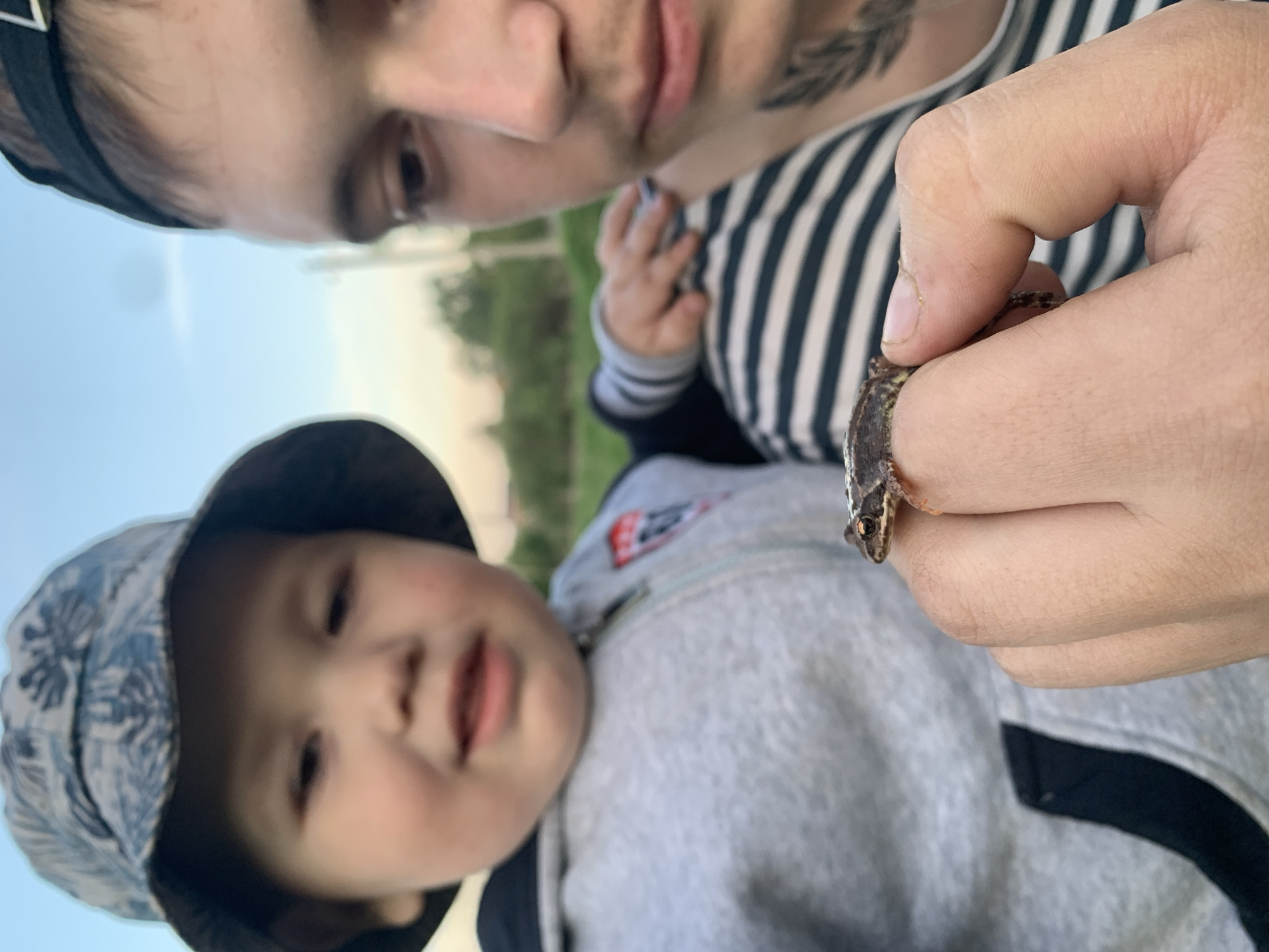 A man and a child are the subjects of an outdoor photograph. The man, wearing a striped shirt and a black headband, is holding a small lizard in his right hand. The child, wearing a blue patterned hat and a gray hooded sweatshirt with black trim, is positioned below and to the left of the man. The background includes a grassy area and a blurred view of trees and sky.
There is no evidence in the image to support that this photo was taken in the city of Фоки, Russia.