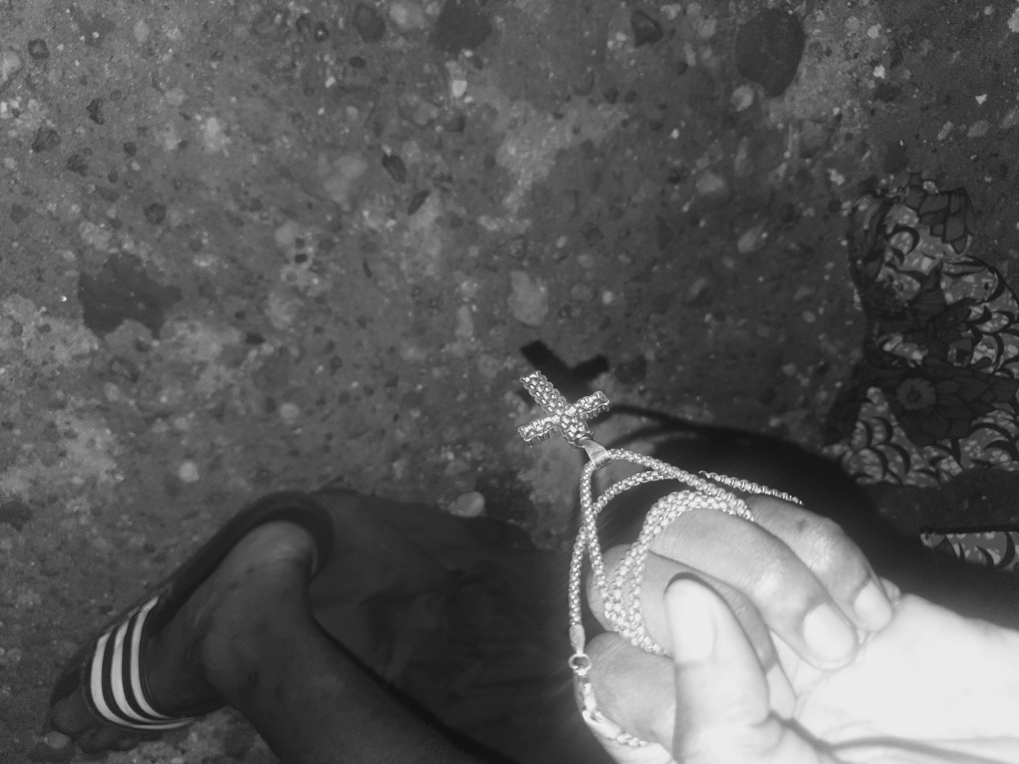 The image, taken in Lokoja, Nigeria, shows a person wearing striped sandals, possibly Adidas, standing on a concrete or rocky surface. The person holds a silver chain with a cross pendant in their right hand. The background includes a textured surface on the left and a patterned cloth or clothing item on the right.