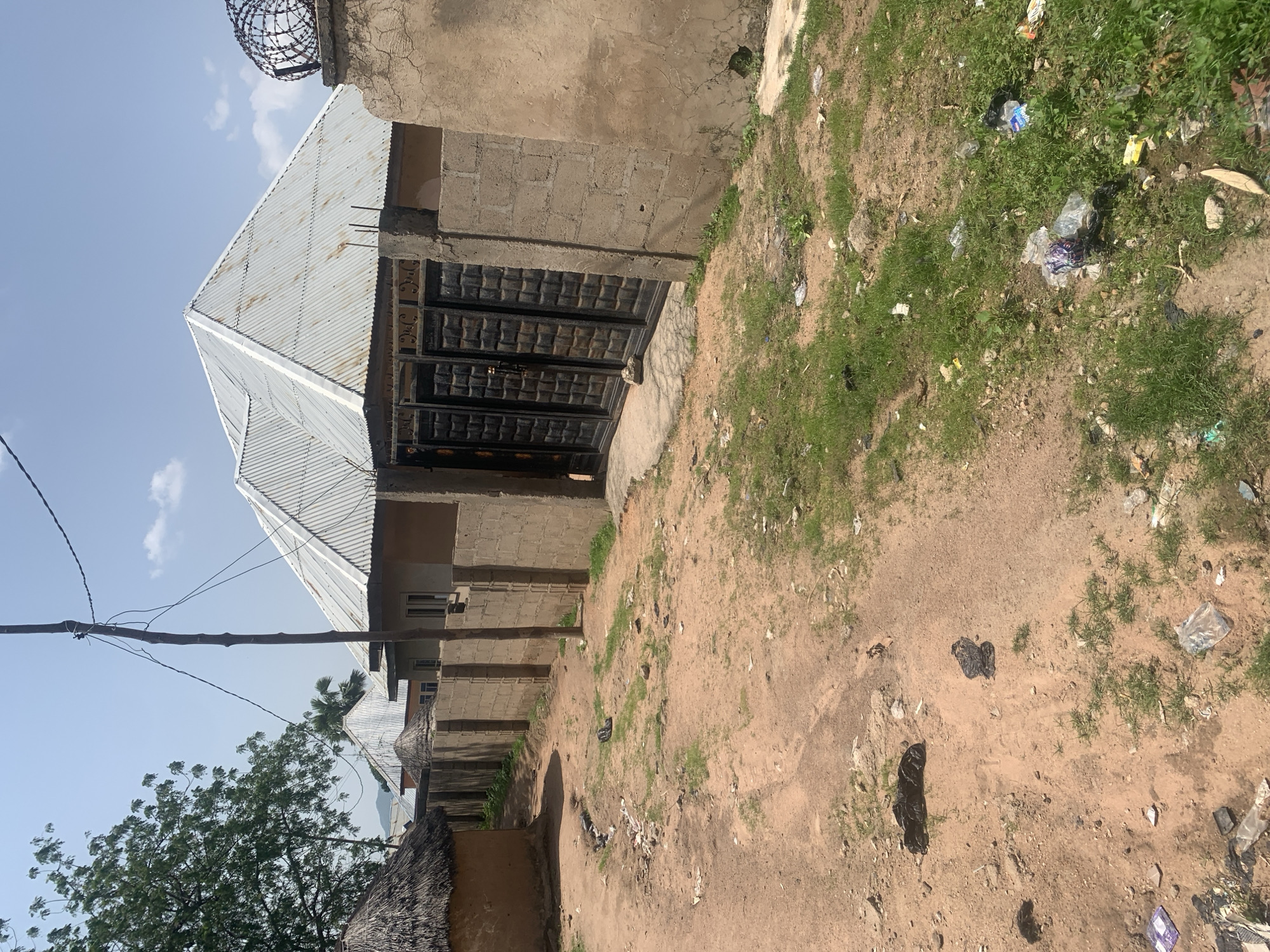 This is a forensic, matter-of-fact description of a scene in Jalingo, Nigeria. The image depicts a dirt road leading to a walled compound. The compound features a modern house with a corrugated metal roof, a black metal gate, and a concrete wall. Adjacent to this structure, there is a traditional hut with a thatched roof. The area is littered with trash, including plastic bags and wrappers, scattered on the ground and in the sparse grass. Electrical wires are visible overhead, supported by a wooden pole. The sky is clear with a few scattered clouds. There are no people visible in the image.