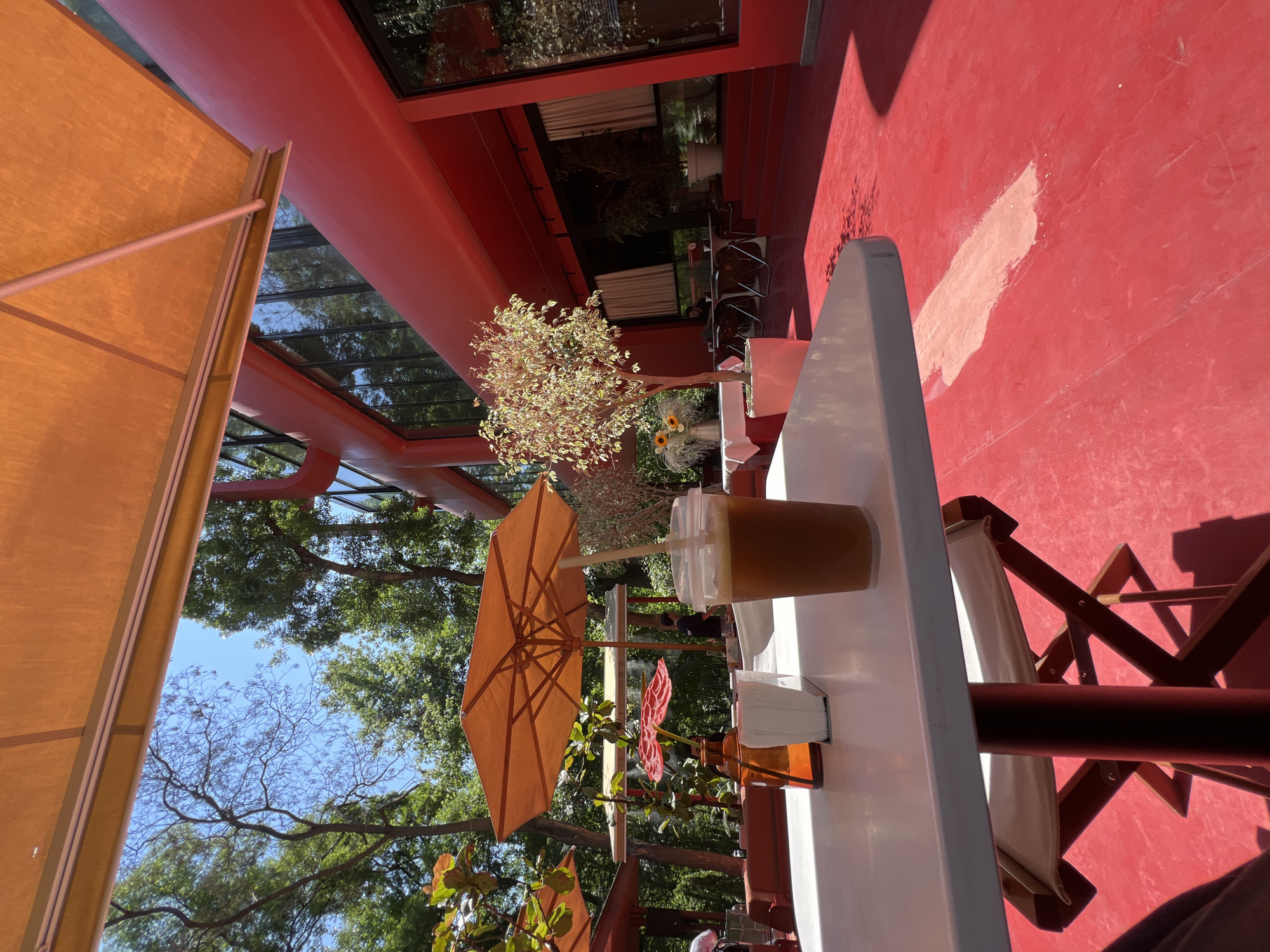 An outdoor seating area with a bright red floor and building. A white table in the foreground holds a clear plastic cup with a brown beverage and a straw, a small orange vase, and a white napkin dispenser. An orange umbrella is angled overhead. In the background, trees and a red modern building with large windows are visible. The location is a cafe in Yerevan, Armenia.