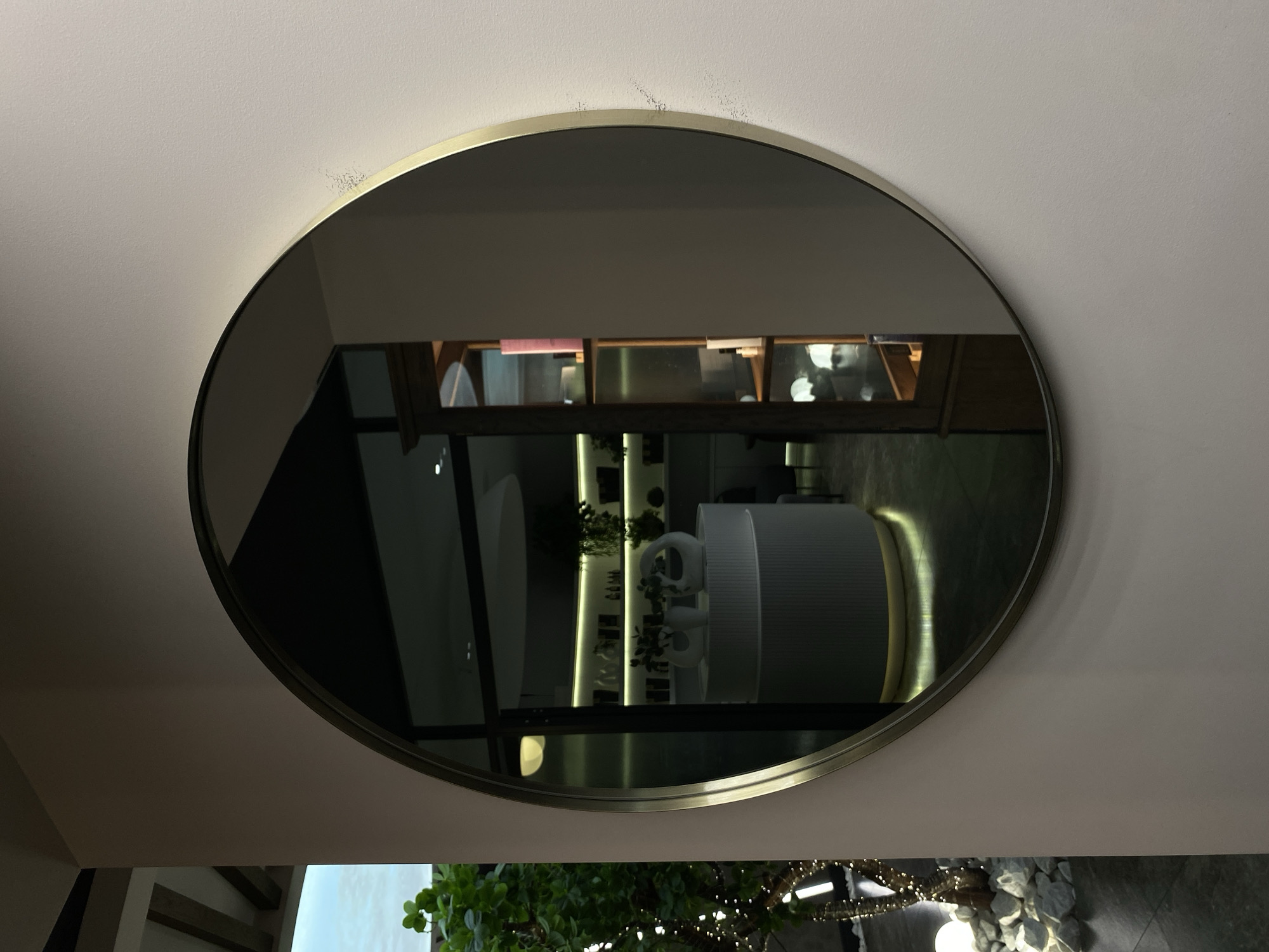 This image shows a round mirror with a thin, metallic frame hanging on a light pink wall. The mirror reflects the interior of a well-lit establishment, possibly a salon or a retail space, with shelves stocked with various products and decorative items. A curved reception desk with vertical detailing and a glowing yellow base is visible in the reflection. To the left of the mirror, a plant with lush green leaves is seen, partially illuminated by string lights. The ground beneath the plant is covered with white stones. The overall scene suggests a modern and chic aesthetic. The location is not identifiable as Votkinsk, Russia.