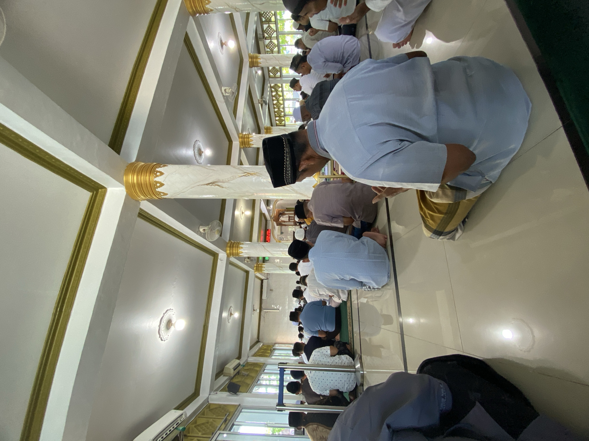 In an interior, likely a mosque, a line of people, predominantly men, are kneeling and bowing, presumably in prayer. Most are wearing light-colored clothing with some wearing traditional Muslim head coverings. The setting is a room with marble-like pillars accented with gold. There are ceiling lights, and a barrier in the foreground that is possibly made of glass or plastic. The location is likely a mosque somewhere in Lhokseumawe, Indonesia.