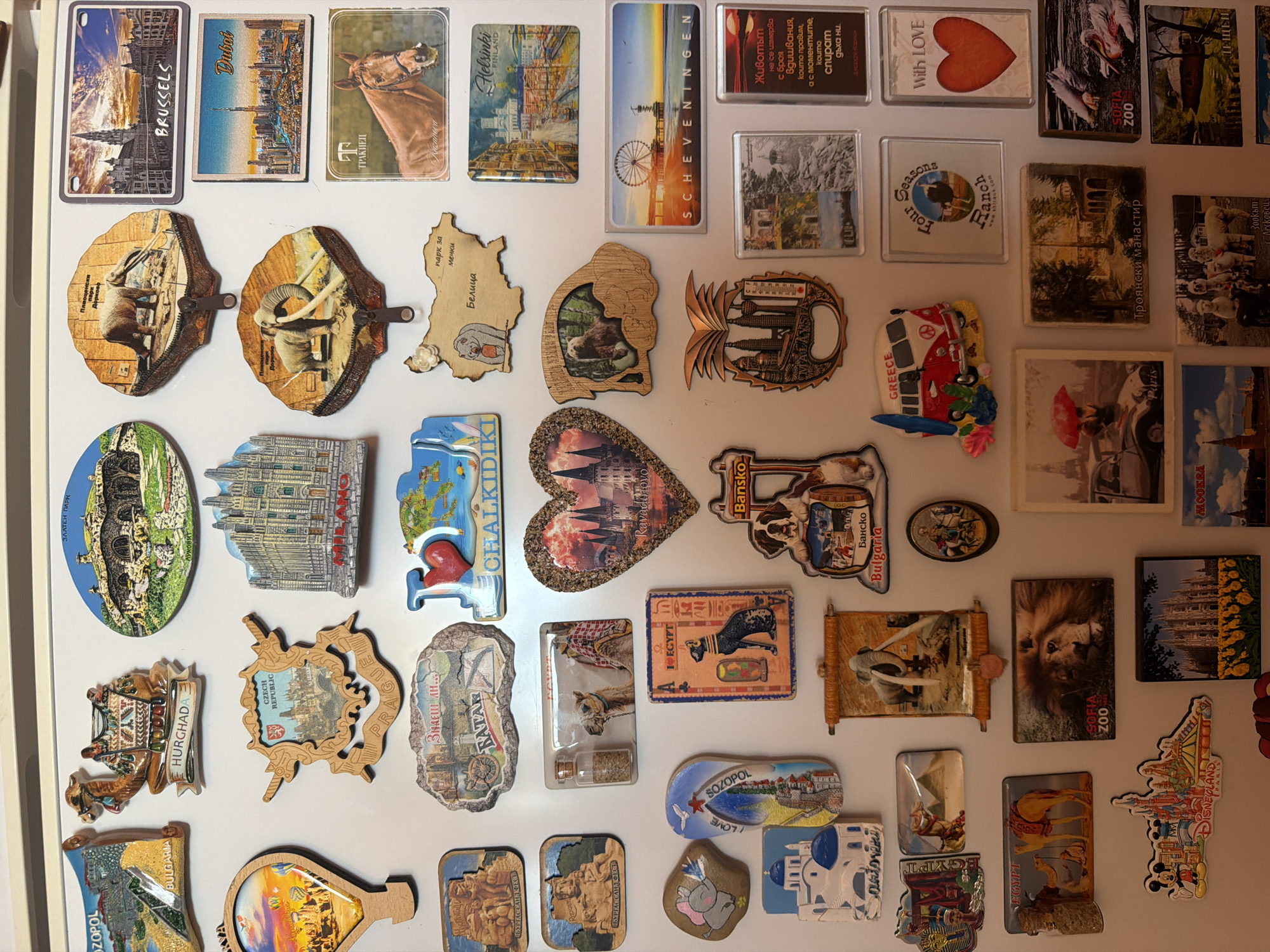 The image is a close-up of a refrigerator door covered in souvenir magnets. The magnets depict various landmarks, animals, and phrases from different places around the world. Cities represented include Brussels, Dubai, Helsinki, Scheveningen, Prague, Paris, and Moscow. There are magnets from Bulgaria, including a Sofia Zoo magnet, and magnets related to Egypt, such as a depiction of an Egyptian cat. Some magnets have wooden frames or accents.