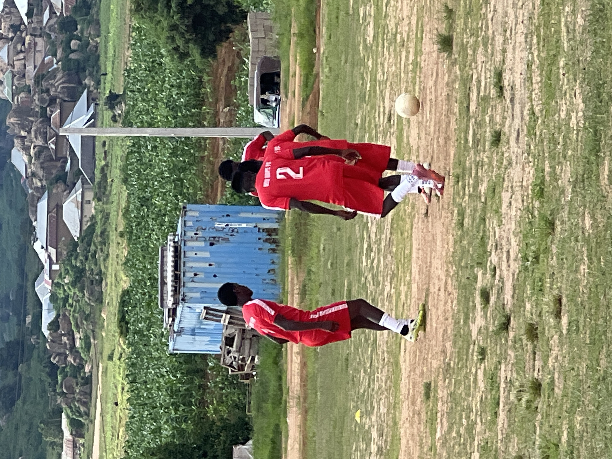 The image depicts a dirt and grass field in Jos, Nigeria, likely a training ground for a local football team. Three young men, dressed in matching red football kits with white accents and trim, are scattered across the field. Two of the men are facing away from the camera, one wearing the number "2" prominently on his jersey. The third player is facing towards the left, also in a red kit. A light-colored football rests on the ground in the foreground, suggesting a pause or an ongoing practice session.
In the background, a large, weathered blue shipping container stands on the left, behind which are piles of wooden debris, possibly remnants of old structures or discarded materials. Beyond the container, a lush green field of crops, likely maize or corn, stretches towards a hilly landscape. The hills are dotted with scattered buildings, some with distinctive grey, angular roofs, interspersed with large rock formations. A white utility pole stands in the middle ground, extending towards the sky. The overall setting suggests a rural or semi-rural environment within or on the outskirts of Jos, characterized by agricultural activity and developing infrastructure. The sky is overcast, indicating a potentially humid or rainy day.