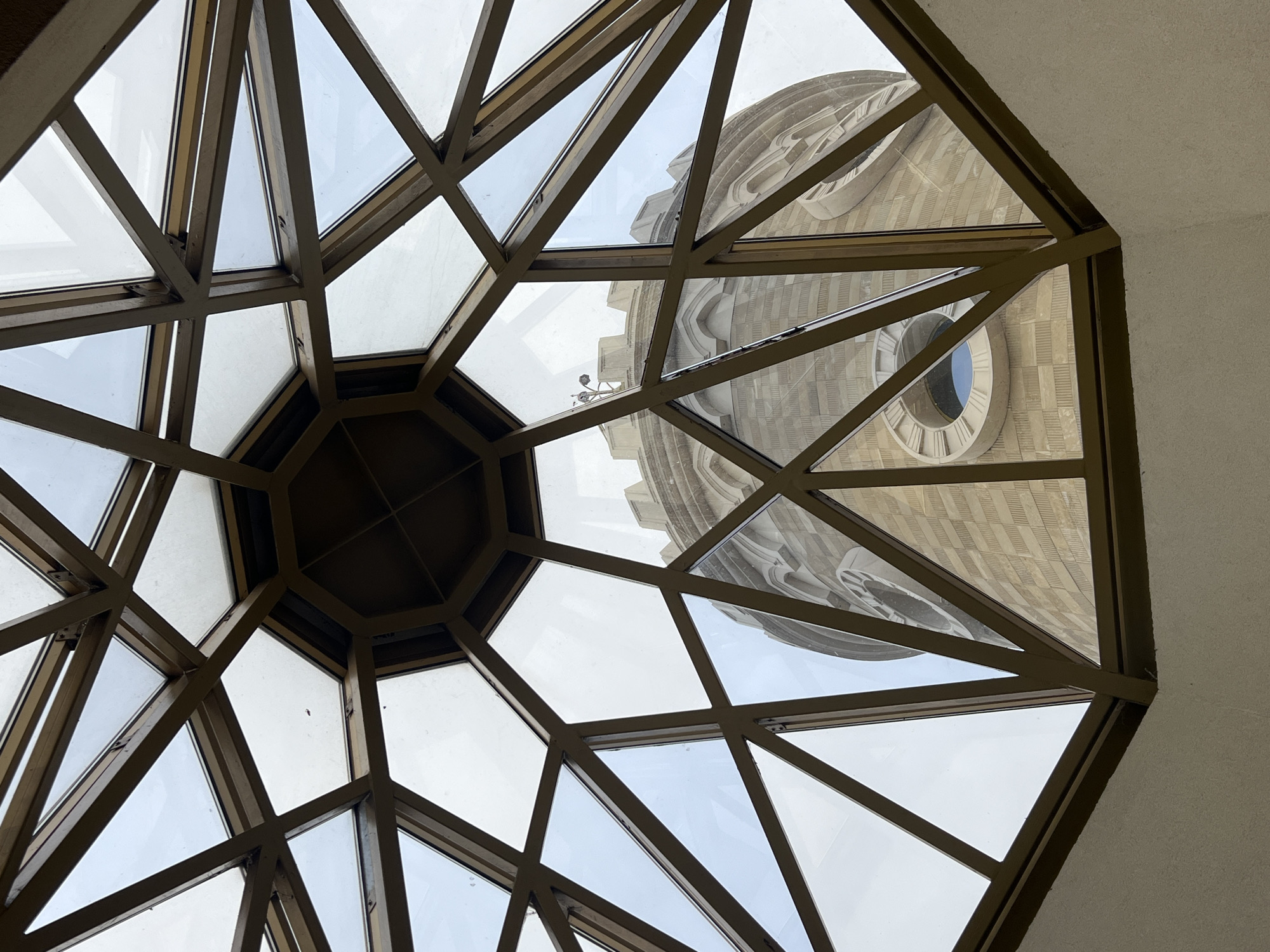 The image shows a view through a geometric glass ceiling, likely a dome, with triangular and trapezoidal panes held by a brown metal frame. Visible through the glass is the exterior of a large, beige stone building featuring a circular window, appearing to be a clock face with Roman numerals. The architecture suggests a religious structure or monument. The location in the city of Kazan, Russia, is incorrect, as the building visible through the glass ceiling appears to be the Sagrada Familia basilica in Barcelona, Spain.