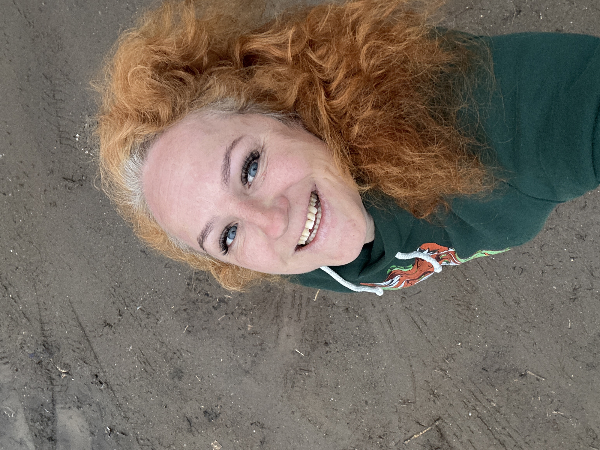 A woman with red curly hair and blue eyes is lying on what appears to be a sandy or dirt-covered surface. She is smiling and wearing a green hoodie. The location appears to be outdoors. The location within the city of Chaikovsky, Russia cannot be determined from the image.