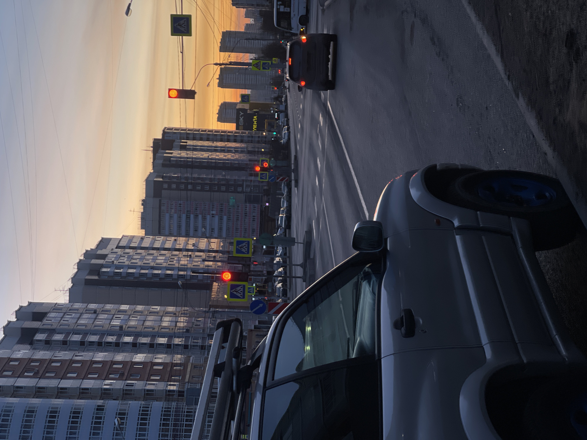 The image depicts a street scene in what appears to be Krasnoyarsk, Russia, recognizable by the "LENTA" store sign in Cyrillic. The view is taken from within a car, with part of the car's body visible in the foreground. Beyond the car, the street stretches into the distance, bordered by tall residential buildings typical of urban Russia. Multiple traffic lights are visible, all showing red. Other vehicles can be seen on the road, including at least one car further down the street. Pedestrian crossing signs are also present. The sky indicates it's either early morning or late evening, with a gradient of colors indicating the sun.