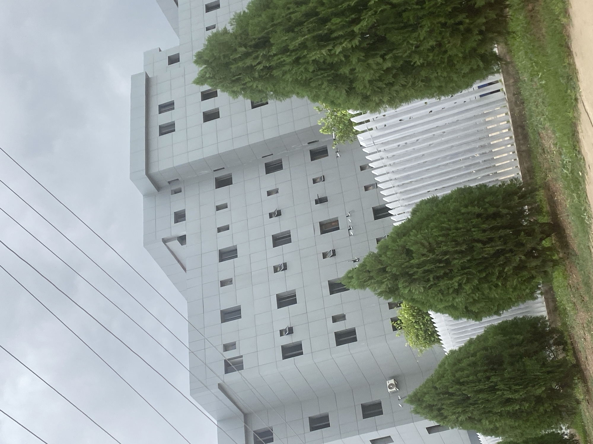 A tall, gray, modern building stands in Lugbe, Nigeria. The building has a grid-like pattern of windows. Power lines run across the left side of the image. Green trees partially obscure the building, and a white fence sits near the trees. Overcast sky.