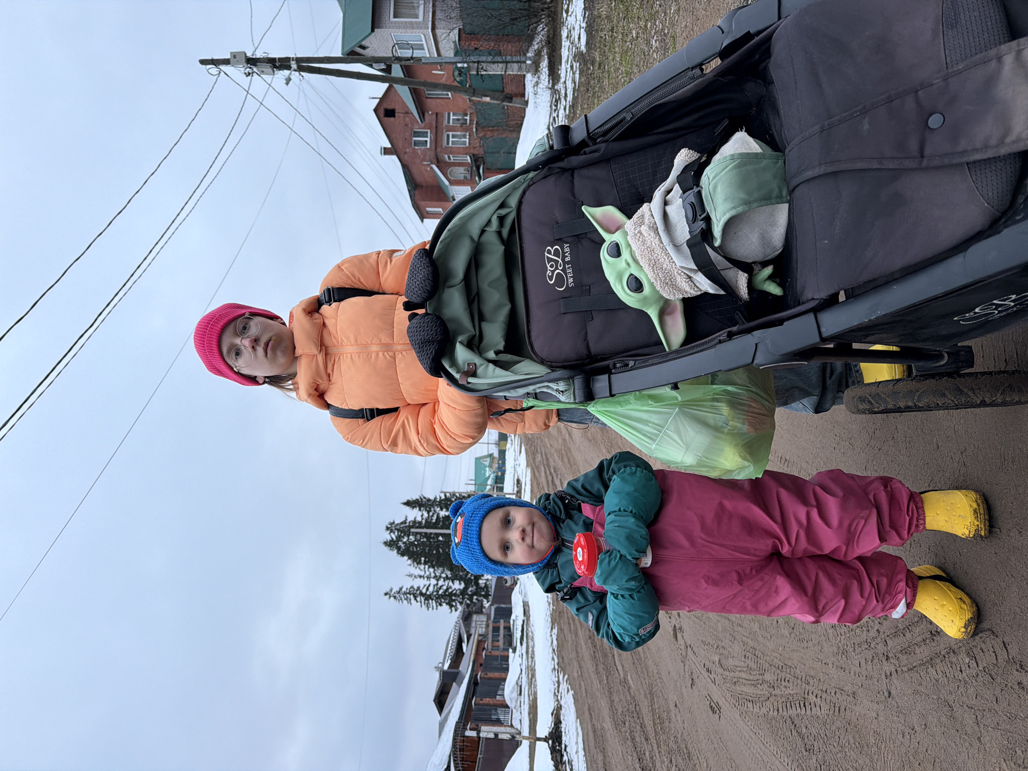 A medium shot shows a person in an orange puffy jacket and pink hat standing beside a black stroller with a Baby Yoda doll inside. The stroller is a "Sweet Baby" brand. A child wearing a blue hat and pink snowsuit stands in front, holding a red container. Both are in an outdoor setting on a dirt road. Buildings and snow patches are visible in the background. The location is determined to be in Chaikovsky, Russia, based on image recognition.