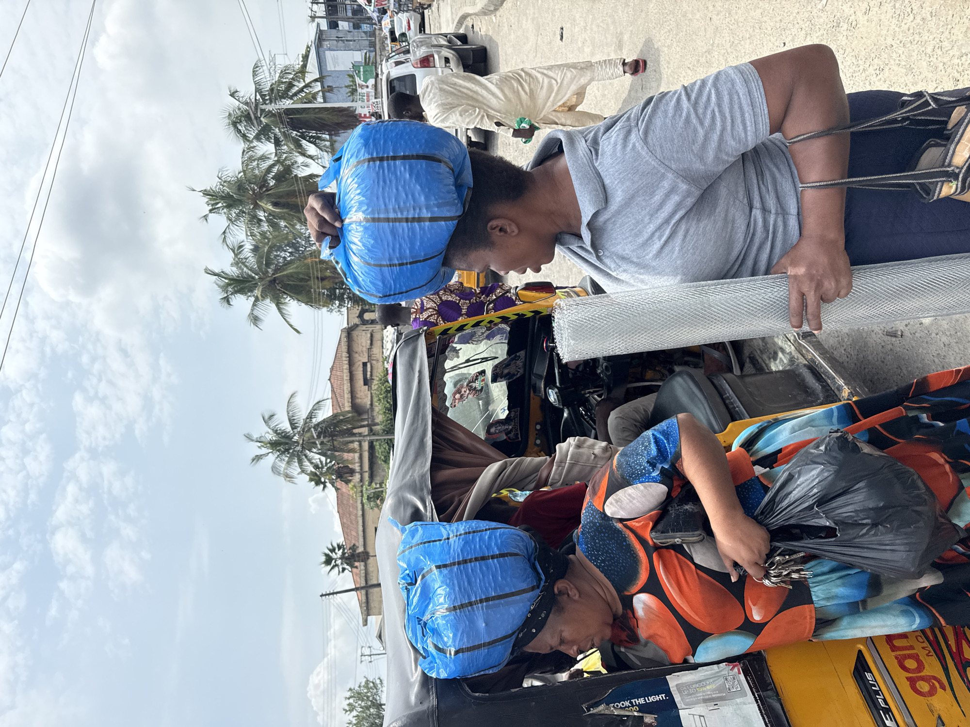 The image shows two women and a man on a street in Ado, Nigeria. A woman in the foreground is carrying a large blue bag on her head and a roll of mesh in her arms. She is wearing a grey shirt and dark blue pants. To her left, another woman, wearing a colorful dress, is also carrying a blue bag on her head and holding a black plastic bag and keys. They appear to be near a yellow auto-rickshaw. In the background, a man in a light-colored robe is walking away, and several vehicles are parked on the side of the road. Palm trees and buildings are visible in the distance, under a cloudy sky. The scene suggests a busy urban environment with people engaged in daily activities.
