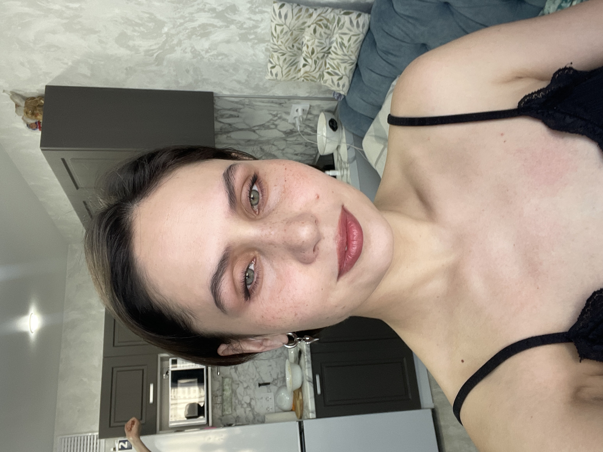 The image is a portrait of a young woman, captured in a selfie-style shot within what appears to be a residential kitchen and living space. She has fair skin with numerous freckles concentrated across her nose and cheeks. Her eyes are a prominent green or hazel, and she has dark, well-defined eyebrows and dark hair that is either short or pulled back. Her lips are adorned with a reddish-mauve lipstick, and she wears a silver-colored hoop earring with a small charm in her visible ear. She is dressed in a black camisole or bralette with thin straps and delicate lace trim.
The background reveals a modern interior. To her left, part of a stainless steel refrigerator and a wall-mounted light fixture are visible. Behind her, a kitchen area features dark grey or brown upper and lower cabinets, with a microwave oven installed above a counter. A white and grey patterned backsplash decorates the wall behind the counter, where a white bowl is also placed. Further back, an unmade bed or sofa with blue upholstery and a cushion featuring a green leaf pattern can be seen, alongside a small white fan. The walls are a light grey with a textured finish. The woman has a calm, direct gaze towards the camera, with a subtle, soft expression. No visible text is present in the image. The scene suggests an everyday moment in a home, located in Izhevsk, Russia.