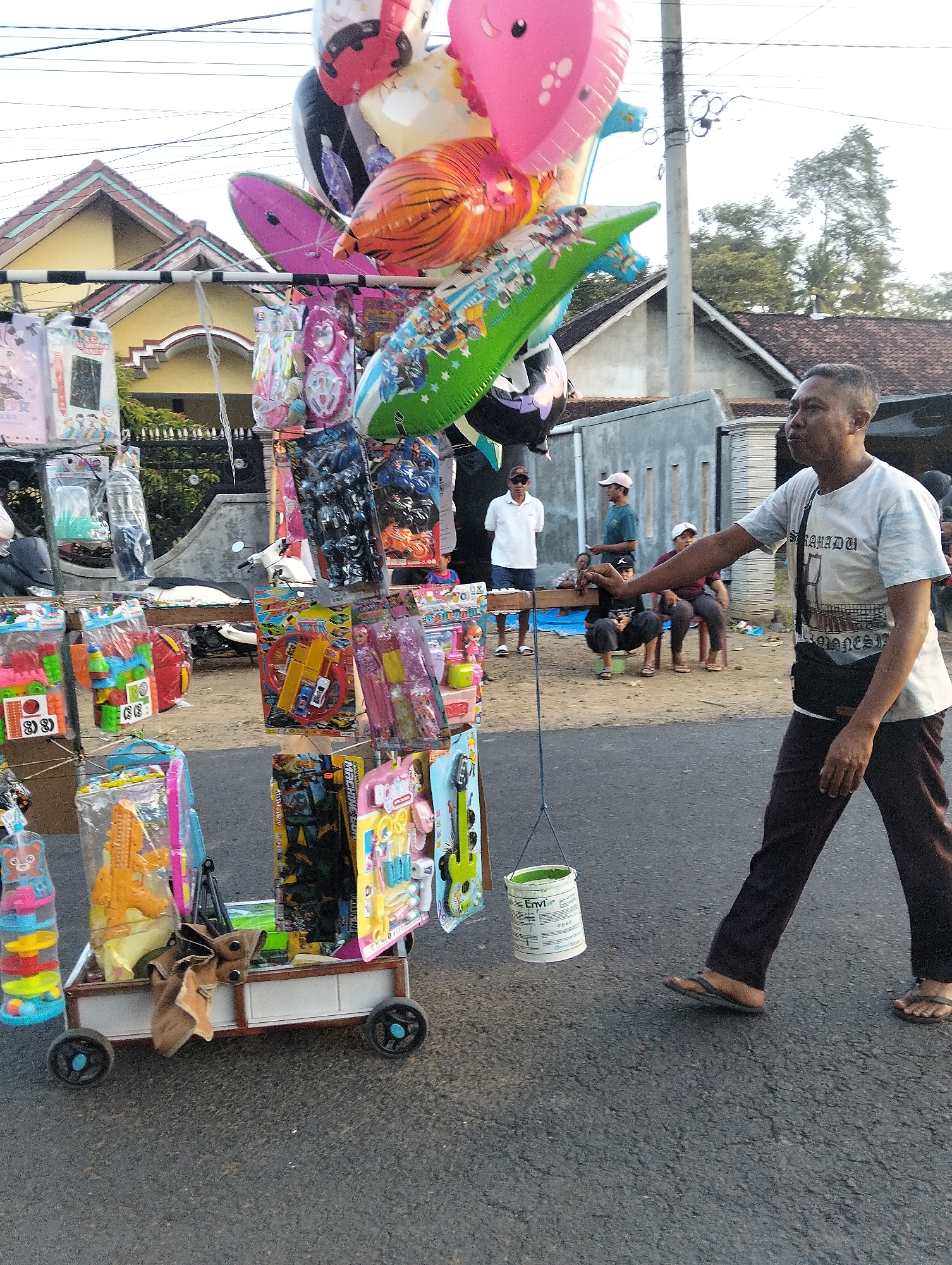 A man is walking across an asphalt road in Sumbersewu, Indonesia, past a mobile stall selling toys and balloons. The man, wearing a white t-shirt and dark pants, has his left arm extended and is looking to his right. The stall is decorated with various colorful balloons and packaged toys, including what appear to be action figures and building blocks. A white bucket is hanging from a string near the stall. In the background, several other people are seated, and houses with tiled roofs are visible. The sky is bright, suggesting it is daytime.