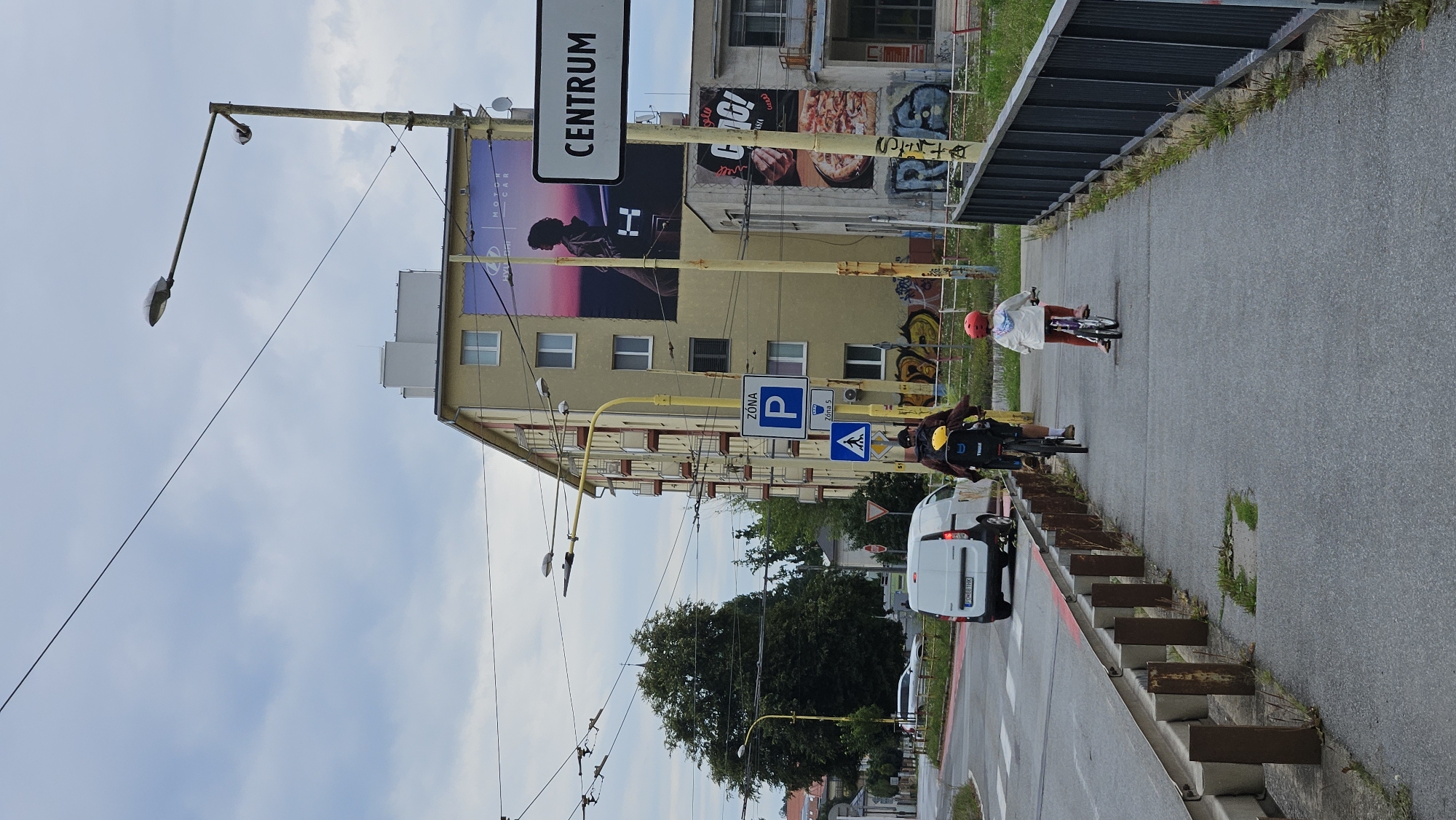 The image shows a street scene in Prešov, Slovakia. In the foreground, two people are riding bicycles on a pedestrian path. The person in front is a child wearing a red helmet and a white jacket, riding a small bicycle. Behind them, an adult wearing a yellow helmet and a backpack is riding a larger bicycle. They are on a paved pedestrian path next to a road with a white van driving away from the camera.
In the background, there is a multi-story yellow building with balconies. A large advertisement featuring a silhouette of a person and the text "CENTRUM" is displayed on the building. Other advertisements and graffiti are also visible on the building's facade. Streetlights and overhead wires are present throughout the scene, indicating an urban environment. The sky is overcast, suggesting cloudy weather. The overall scene depicts an ordinary day in a Slovakian city with people engaged in recreational activities.