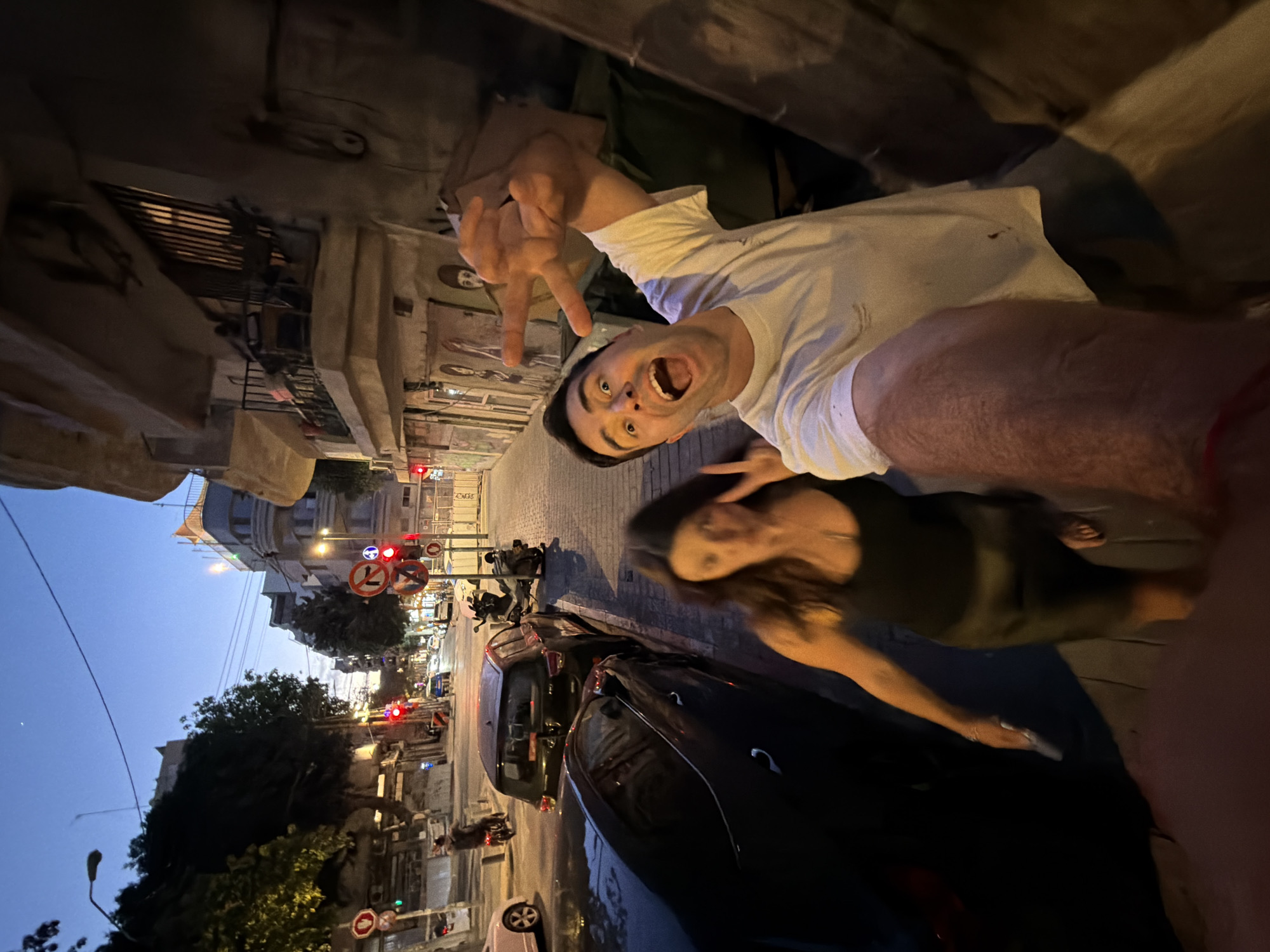 A medium-shot, low-angle, portrait-oriented photograph captures two people posing in a Tel-Aviv, Israel street. A man in a white T-shirt, making a peace sign, has his mouth open in a shout. A woman stands to his right, slightly blurred, and appears to be holding a cell phone.
The background shows a streetscape at dusk. Cars are parked along the street, and traffic signals are visible in the distance. Street signs with Hebrew characters and European traffic signs are present. The buildings lining the street are of light-colored stone with street art.