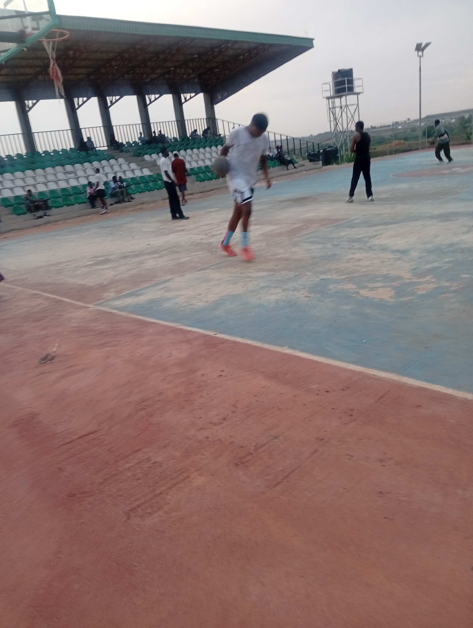 In an outdoor basketball court in Jos, Nigeria, a young man in a white t-shirt and shorts dribbles a basketball. He is mid-action, looking down at the ball. The court is a light blue and white, with a red dirt perimeter. In the background, a covered grandstand with green and white seats is visible. Several people are seated in the grandstand and some are standing on the court. A water tower and a light pole stand on the right side of the court. The sky is overcast.
