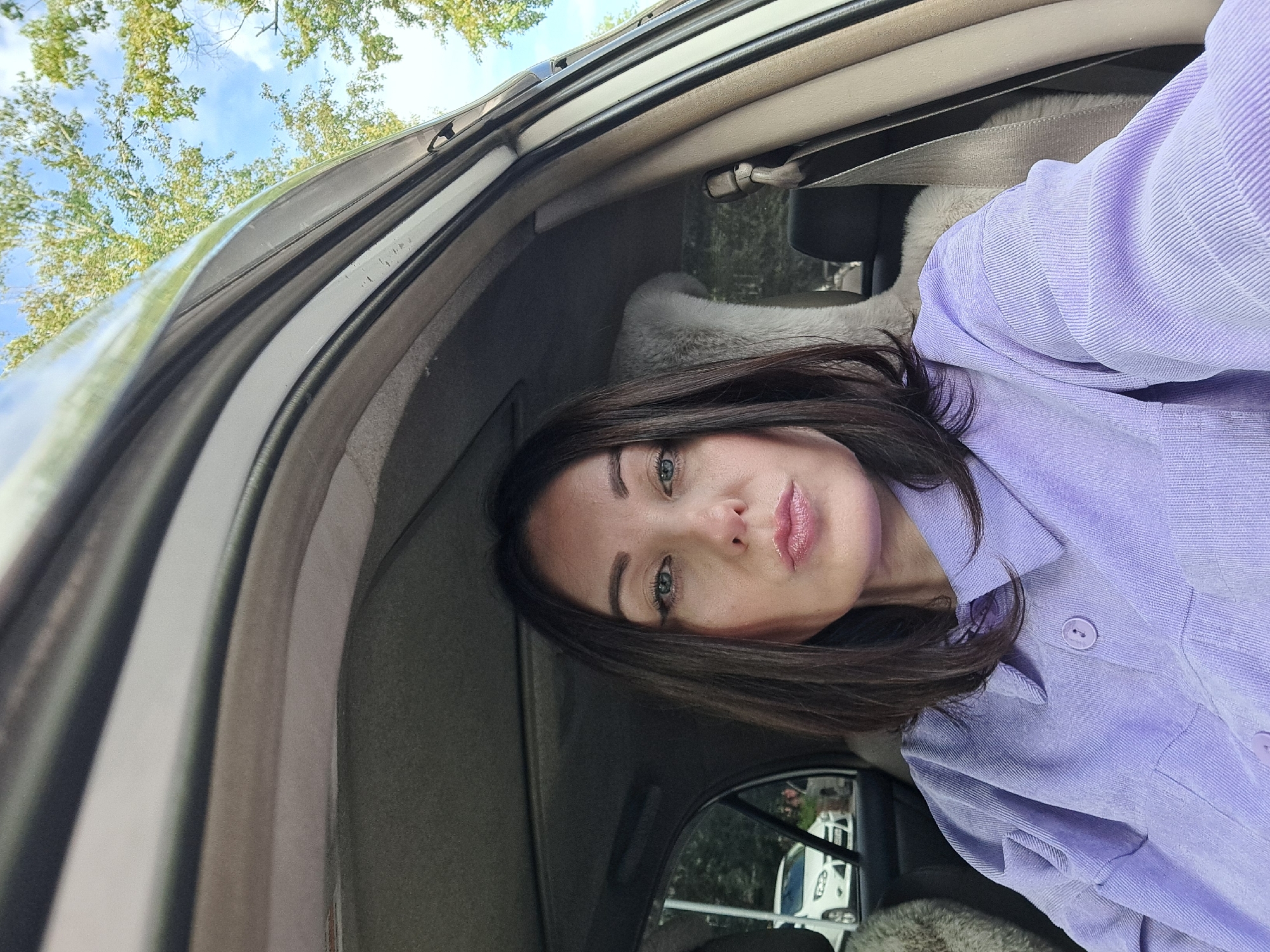In this close-up, medium shot, a woman with dark brown hair and blue eyes is taking a selfie from the driver's seat of a car. She is wearing a light purple corduroy button-up shirt. Her mouth is slightly puckered, and her gaze is directed at the camera. The interior of the car is visible, including the car's ceiling, a tan colored seatbelt, and a furry seat cover. Through the car window on the left, a white car is visible parked on a street, suggesting an urban or suburban setting. Trees with green and yellow leaves are visible outside the car, indicating an autumn season. The sky is blue with some scattered clouds. The context of Irkutsk, Russia is not discernable from the visual elements of the image.