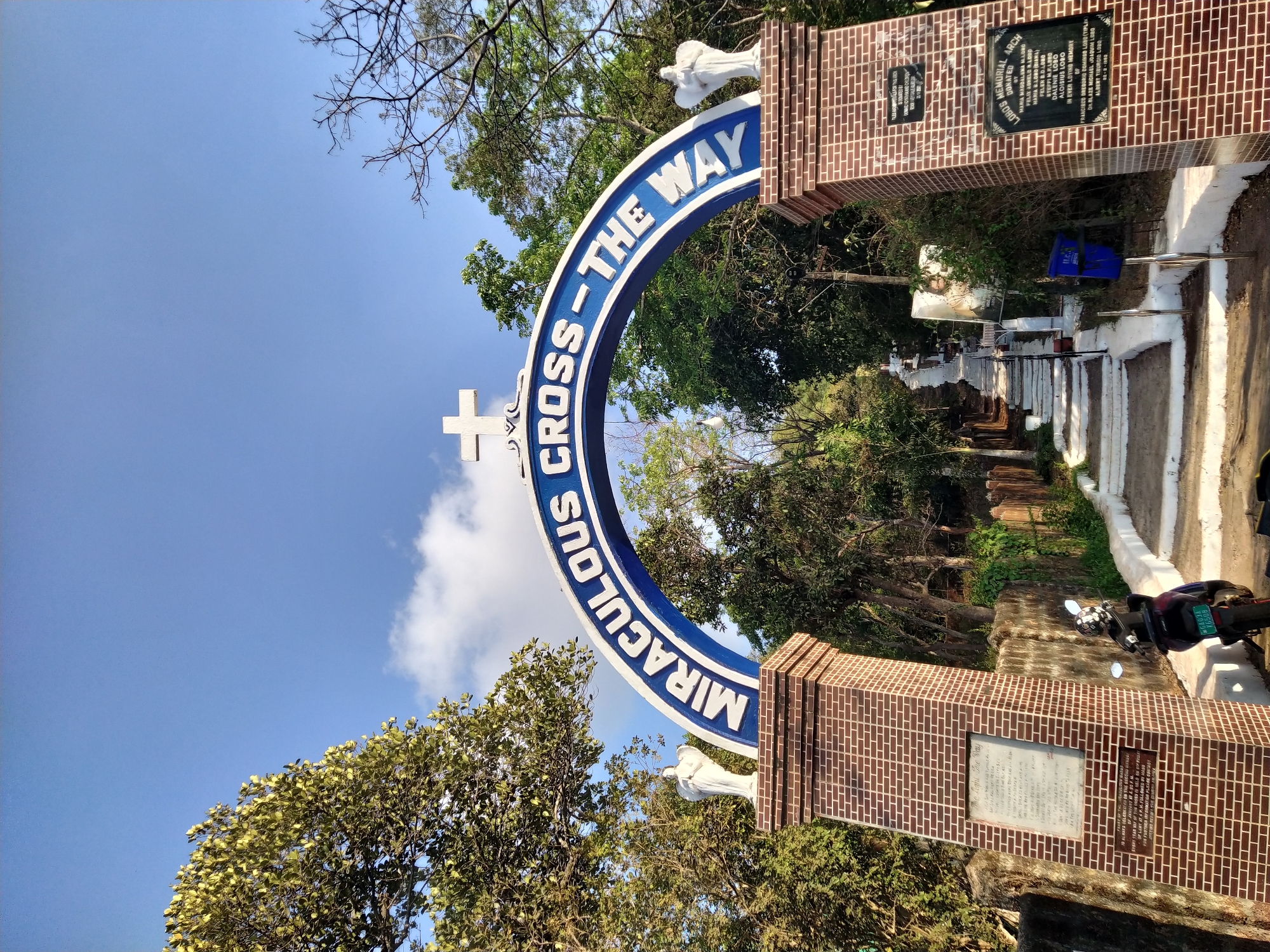 The image shows the entrance to "Miraculous Cross-The Way," located in Assagao, India, as noted by the "Lobo's Memorial Arch" plaque visible. The entrance consists of a semi-circular blue sign with white lettering supported by two brick pillars. A white angel statue adorns the top of each pillar. The sign spans a pathway leading into a cemetery or memorial garden. Plaques are affixed to each pillar with memorial inscriptions. A motorcycle is parked nearby. The sky is blue with some clouds and trees are visible around the perimeter.
