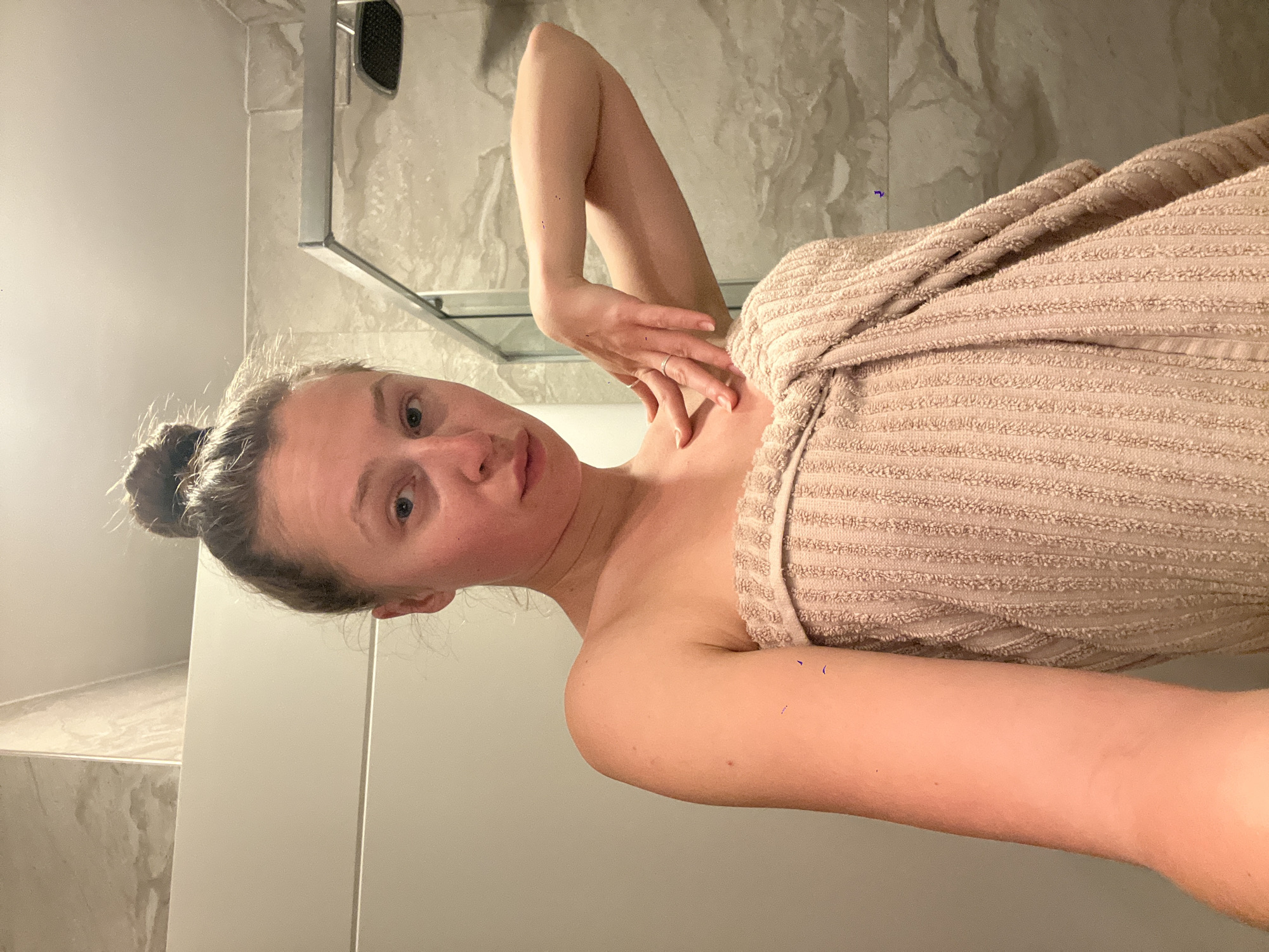 A woman stands in a bathroom, wrapped in a beige towel, with her hair pulled up in a bun. She is holding her arm up, with her hand near her neck. The bathroom appears to have a modern design with marble-like walls, a shower enclosure with a silver frame, and a white cabinet. The location is not discernable in the photograph.