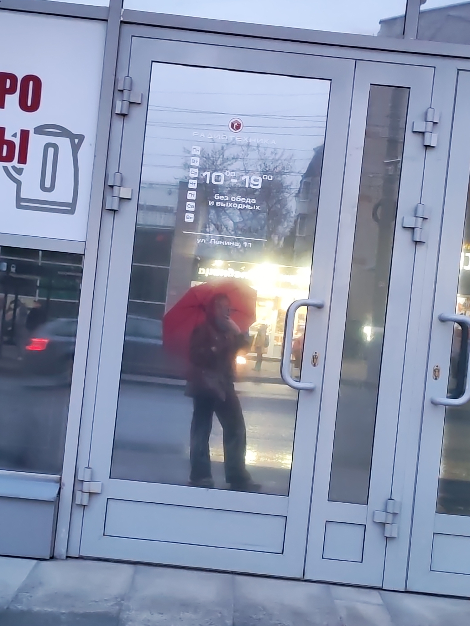 The image depicts a storefront in Izhevsk, Russia, specifically at 11 Lenina Street, as indicated by the text on the storefront's window. The store's sign features the words "РАДИОТЕХНИКА" (Radio Equipment), along with the store's operating hours, which are from 10:00 to 19:00, and it is open every day.
A person holding a red umbrella is visible through the storefront's glass doors. The storefront itself is a modern construction, with white frames and glass panels. The surrounding environment appears to be urban, with street-level activity reflected in the glass. The lighting suggests it is either early morning or late afternoon.