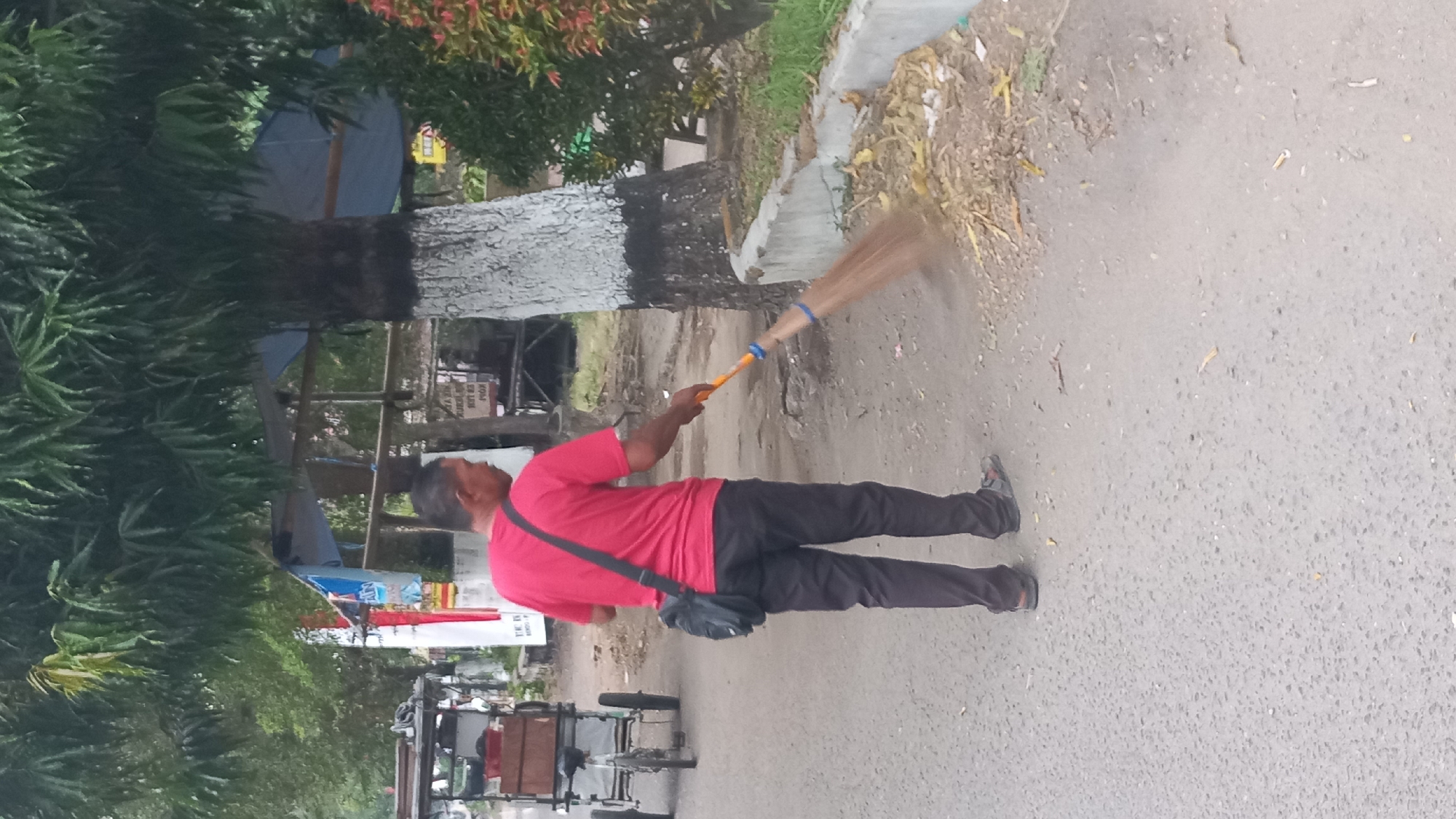 A man in a red t-shirt and black pants is sweeping leaves with a broom on the side of a paved road in Pare, Indonesia. A cart with wheels and wooden crates is parked to the left of the man. Trees with white-painted bases line the road. The ground is covered with scattered leaves and debris.
