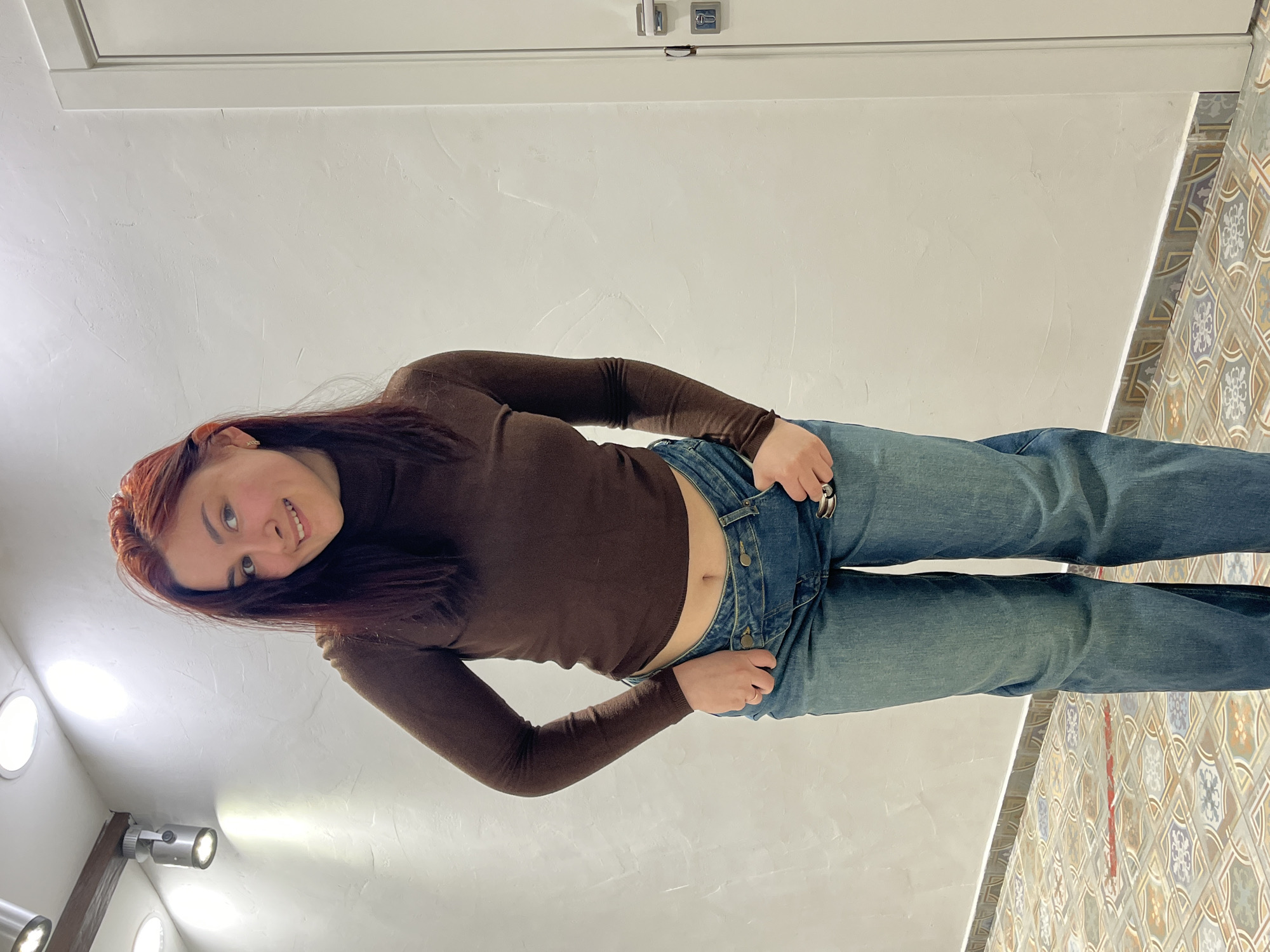 A female subject stands centrally in an indoor setting in Neftekamsk, Russia. She has reddish-brown hair and a slight smile, looking directly at the camera. She wears a dark brown, long-sleeved, cropped turtleneck shirt, exposing her navel, and light blue denim jeans with a distinctive overlapping waistband design. A silver-colored ring is present on her right ring finger. The background features a textured, light-colored wall with recessed circular ceiling lights and adjustable spotlights mounted on a wooden beam, providing artificial illumination. A white door with a silver handle and lock mechanism is visible on the right side. The floor is covered with multi-colored patterned tiles, separated from the wall by a grey-white baseboard. A thin red line is visible on the floor in the lower-left quadrant.