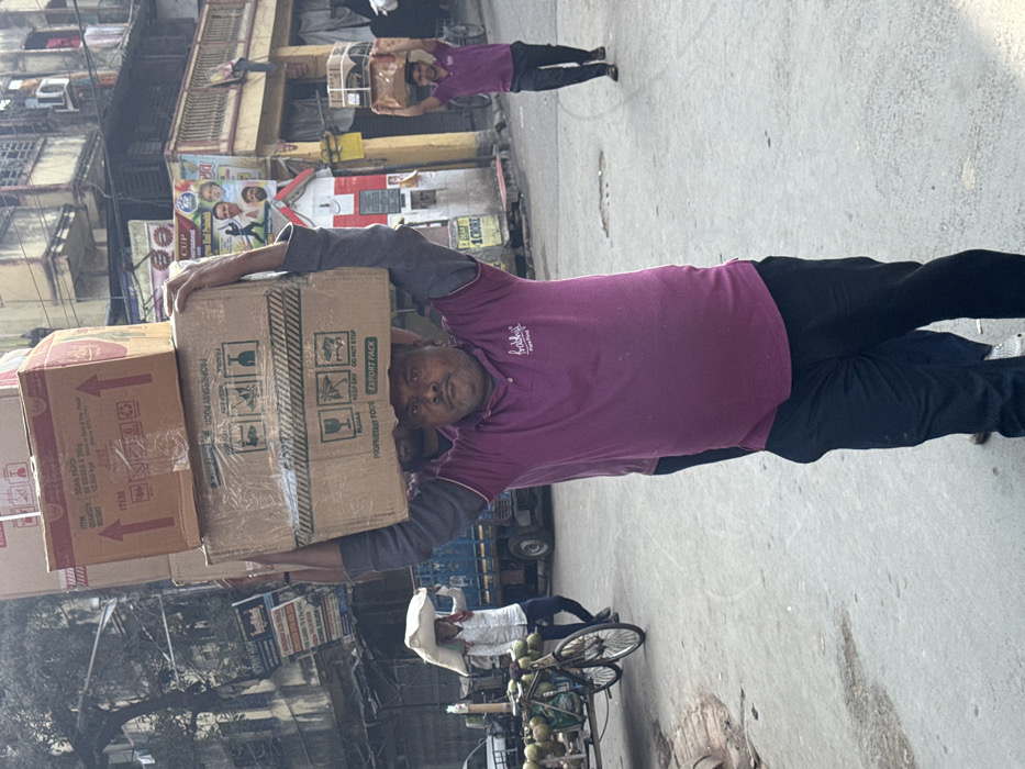 A man in a purple t-shirt balances a stack of cardboard boxes on his head on a city street. Text on the boxes reads "PROPRIETARY FOOD" and "EXPORT PACK." Behind him, another man balances boxes on his head in the distance. A coconut vendor sits on a bicycle cart laden with coconuts. Buildings and billboards line the street. A billboard advertises "1 CRORE".