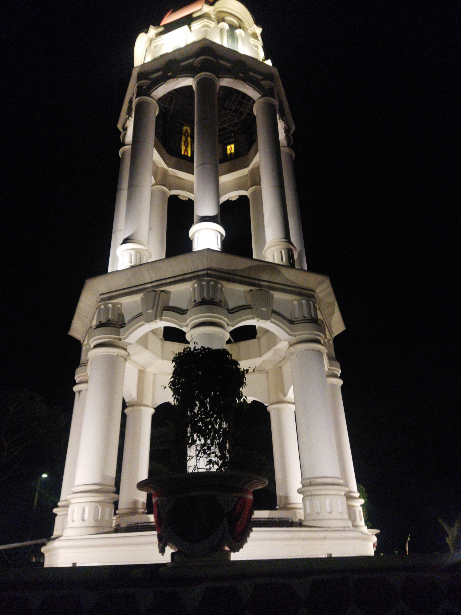 The image shows a close-up, low-angle shot of the Tugu Muda monument in Semarang, Indonesia at night. The white concrete monument is illuminated by artificial lights, highlighting its classical architectural features. The monument has a multi-tiered structure with prominent columns and ornate detailing. In the foreground, a dark, leafy plant in a large planter is visible, partially obscuring the base of the monument. The background is a stark black sky, devoid of any celestial bodies or other distractions. The lighting creates strong shadows and emphasizes the monolithic form of the monument.