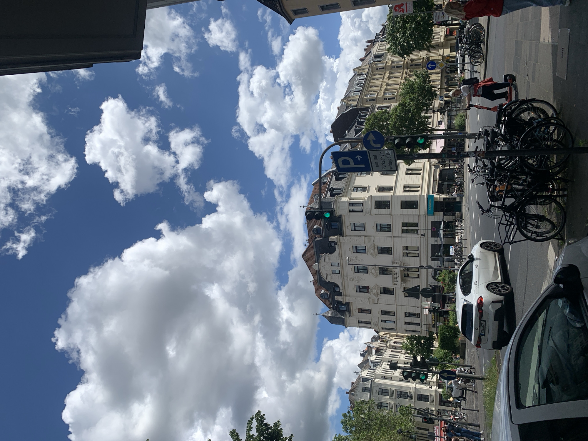 The image, rotated 90 degrees counter-clockwise, captures a vibrant urban street scene under a bright, sunny sky. The sky, dominant on the left side of the rotated image, is a vivid blue filled with numerous large, fluffy white cumulus clouds.
The scene transitions to the right where a row of multi-story buildings with detailed, light-colored facades and darker sloped roofs line a street. Green trees are scattered along the sidewalk, adding foliage to the urban landscape.
At street level, several subjects and activities are observable. Multiple bicycles are parked in metal racks along the sidewalk. A traffic light, displaying a green signal, indicates an active intersection or crossing. Prominently displayed is a blue parking sign featuring a 'P' symbol, an upward arrow, and the German text "nur mit Parkschein werktags 9-20h" (only with parking ticket weekdays 9 AM - 8 PM). Another blue circular sign with a white arrow pointing right is also visible.
Several people populate the scene. One individual in a red jacket and light pants stands near a building entrance on the sidewalk. Another person, wearing a white t-shirt with red stripes and dark pants, is actively riding an orange/red electric scooter along the sidewalk. More people can be seen in the background, walking or standing.
Vehicles include a white car, likely a hatchback, parked on the street, displaying the German license plate "KL E 575". In the extreme foreground on the right, parts of a gray or silver car, including its hood, windshield, and side mirror, are visible. On a building in the background, text "AGNES+" is faintly discernible.
The bright daylight, clear weather, and specific German text on the parking sign and car license plate confirm the setting is a German city during daytime, likely on a weekday.