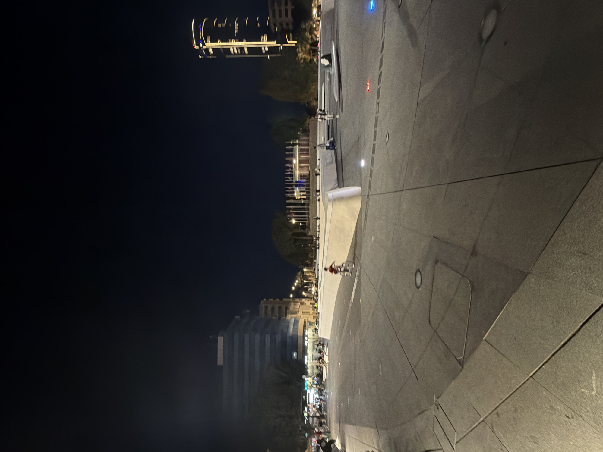 The image depicts an urban plaza at night in Nicosia, Cyprus. A tall, modern skyscraper with illuminated vertical lines is visible in the background to the right. To the left, a multi-story building with horizontal windows and a darker facade dominates the mid-ground. In the foreground, a large, paved plaza area with geometric patterns and embedded lights is visible. A person is seen riding a bicycle in the plaza, moving from right to left. Several other individuals are present in the background, some walking and others gathered in small groups. A line of flags is visible further in the background, in front of a row of lights. The overall scene suggests a public space used for recreation and social gathering in the evening.