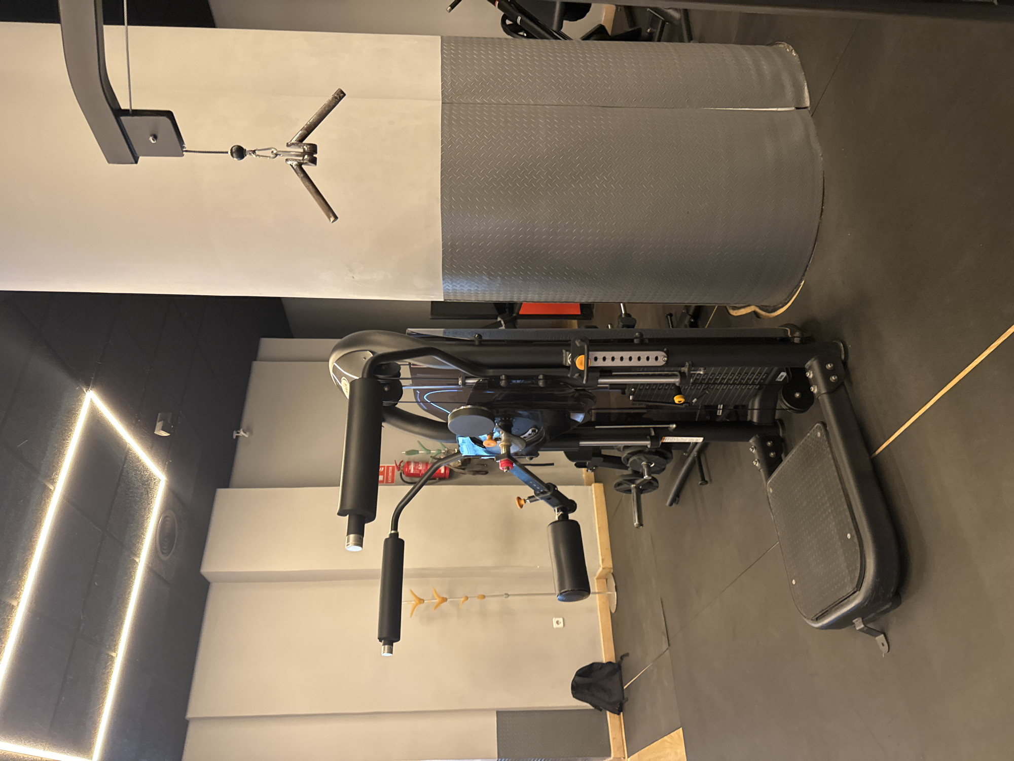 The image shows a gym in Murcia, Spain, featuring a multi-station weight machine with various attachments. The machine has padded arm rests and a weight stack. To the left of the machine, a black backpack rests on the black rubber gym floor. A coat rack with orange hangers stands against a light gray wall. In the background, a red fire extinguisher is mounted on the wall. A large, light gray cylindrical pillar with a diamond-plate textured lower section is to the right of the machine. Above, a rectangular LED light fixture illuminates the ceiling. A single cable with a V-bar attachment hangs from a pulley system attached to the pillar.