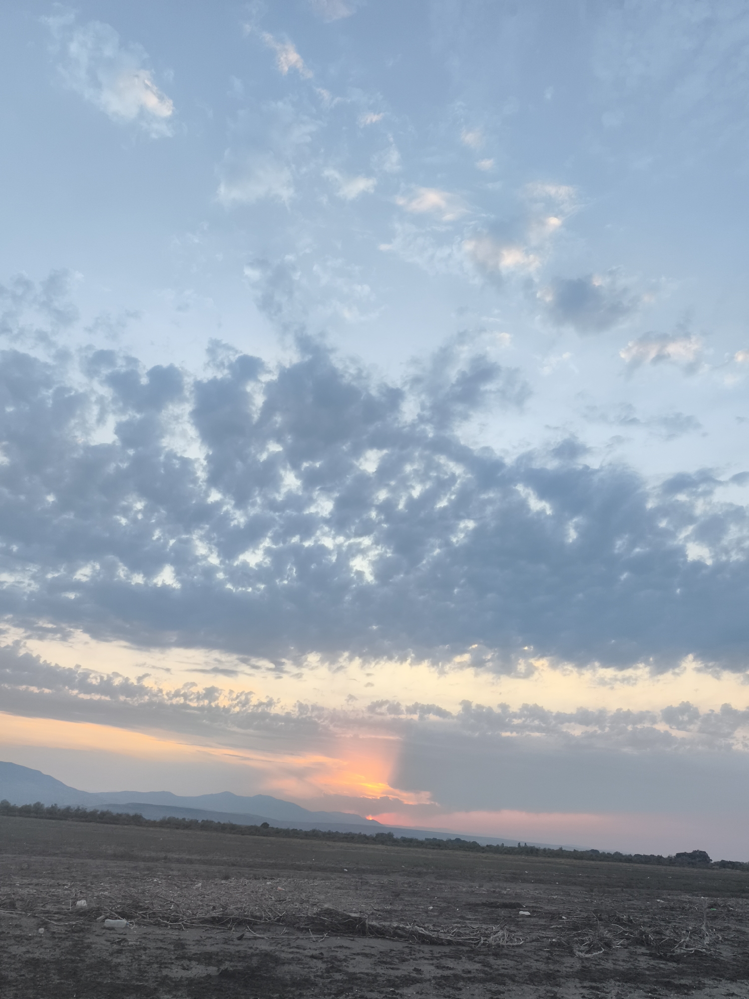The image depicts a wide shot of a rural landscape at dusk, with the sun setting behind distant mountains. The sky is filled with a mix of grey and white clouds, with streaks of orange and pink near the horizon. In the foreground, there is a dirt field with some scattered debris. The overall scene is serene and peaceful, indicative of a rural setting, possibly in the outskirts of Balaca Həmyə, Azerbaijan, given the mountainous terrain. There are no people or animals visible in the frame.