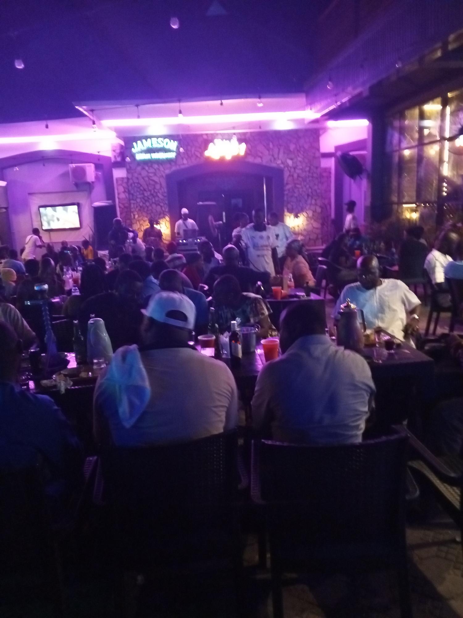 This image depicts an indoor scene at a bustling bar or lounge, likely in Lokoja, Nigeria, during the nighttime. The venue is bathed in vibrant purple and pink neon lighting, creating a lively and contemporary atmosphere.
Numerous people, predominantly men, are visible throughout the space. Many are seated at dark tables, facing away or partially towards the camera, engaged in socializing and drinking. Some individuals are standing, particularly in the background near an elevated area that appears to be a stage or DJ booth. On this elevated platform, a person in a white shirt is visible alongside equipment, suggesting a DJ or live entertainment.
Visible objects on the tables include various bottles, glasses, and at least one shisha pipe. In the background, a flat-screen television is mounted on the wall, displaying content. An air conditioning unit is also visible. The walls behind the central area feature a textured, possibly stone, finish.
A prominent neon sign on the back wall clearly displays the text "JAMESON" in green, indicating a brand presence. Below this, additional, less distinct text is present. The overall scene conveys a social gathering, with patrons enjoying drinks and entertainment in a dimly lit, energetic environment.