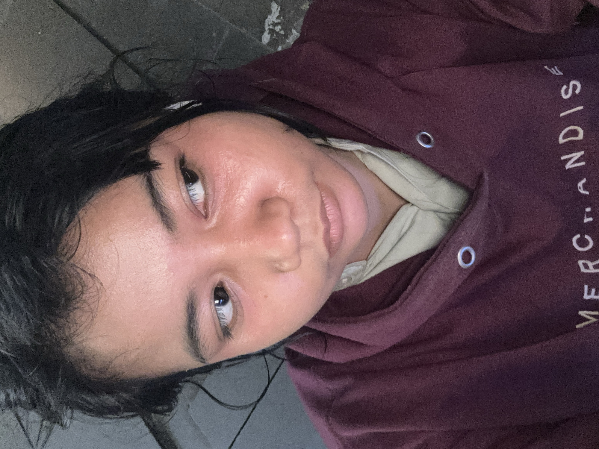 The image is a close-up, selfie-style photograph of a young person wearing a maroon hooded sweatshirt over a cream-colored shirt. The person has dark, messy hair and their face is illuminated with a reddish hue, possibly from lighting or a skin reaction. Their eyes are looking to the right, and their lips are slightly parted. The background appears to be an outdoor urban setting, with a gray, textured surface that could be a wall or pavement. Part of the hooded sweatshirt reveals white text that reads "MERCHANDISE". While the specific location of North Jakarta, Indonesia, is provided, the visual information within the image itself does not offer any direct clues to confirm this specific geographical context.